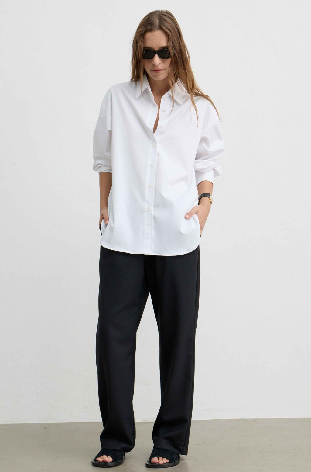 A woman modeling an oversized white button-down shirt with black wide-leg trousers and black sandals, standing against a plain white studio wall.