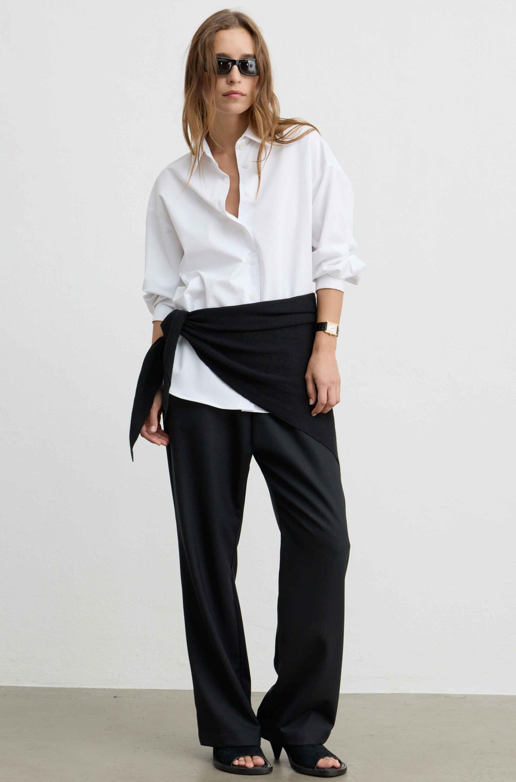 A model wearing a white button-down shirt, black wide-leg trousers, and black sandals, styled with a black fabric wrap tied around the waist, set against a plain light gray studio background.