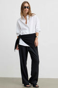 A model wearing a white button-down shirt, black wide-leg trousers, and black sandals, styled with a black fabric wrap tied around the waist, set against a plain light gray studio background.