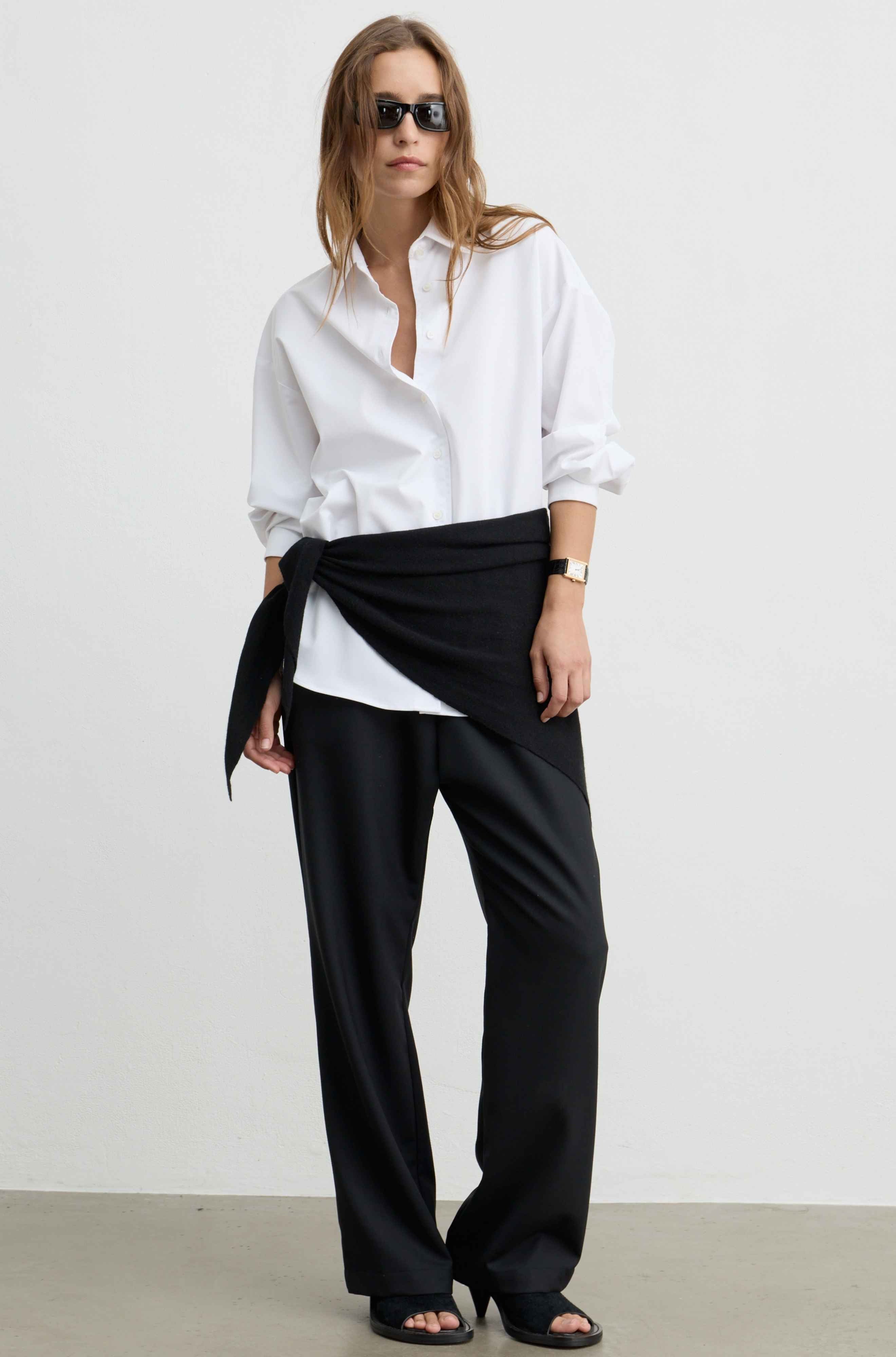 A model wearing a white button-down shirt, black wide-leg trousers, and black sandals, styled with a black fabric wrap tied around the waist, set against a plain light gray studio background.