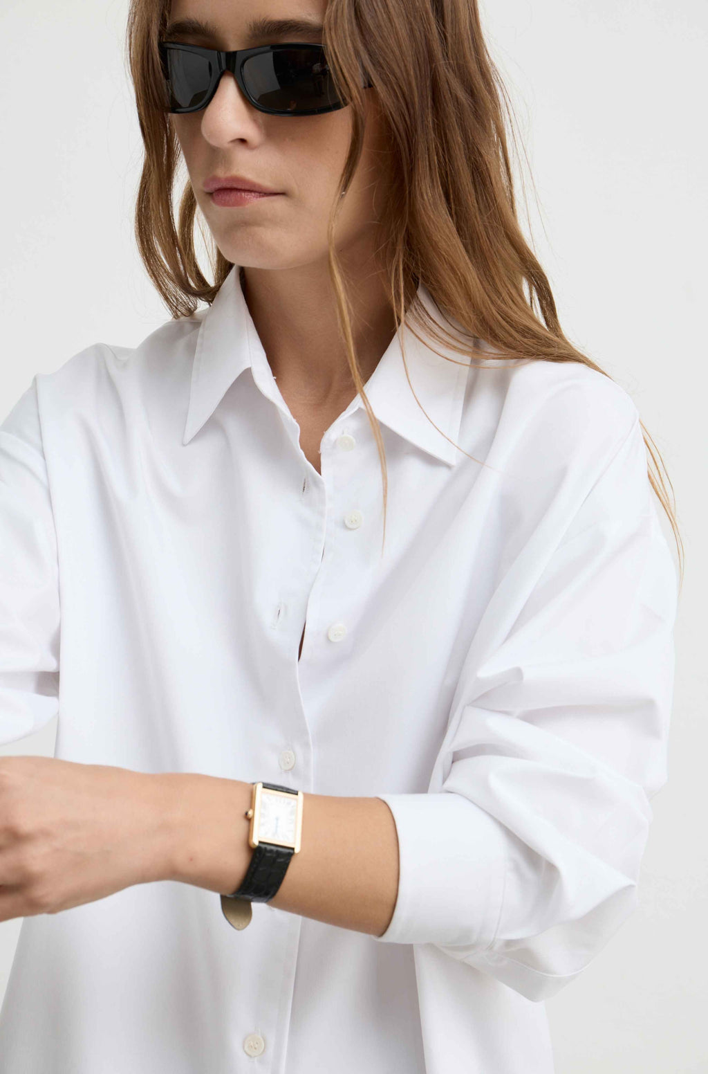 A woman wearing an oversized white button-down shirt and black sunglasses, shown from the chest up against a plain light background.