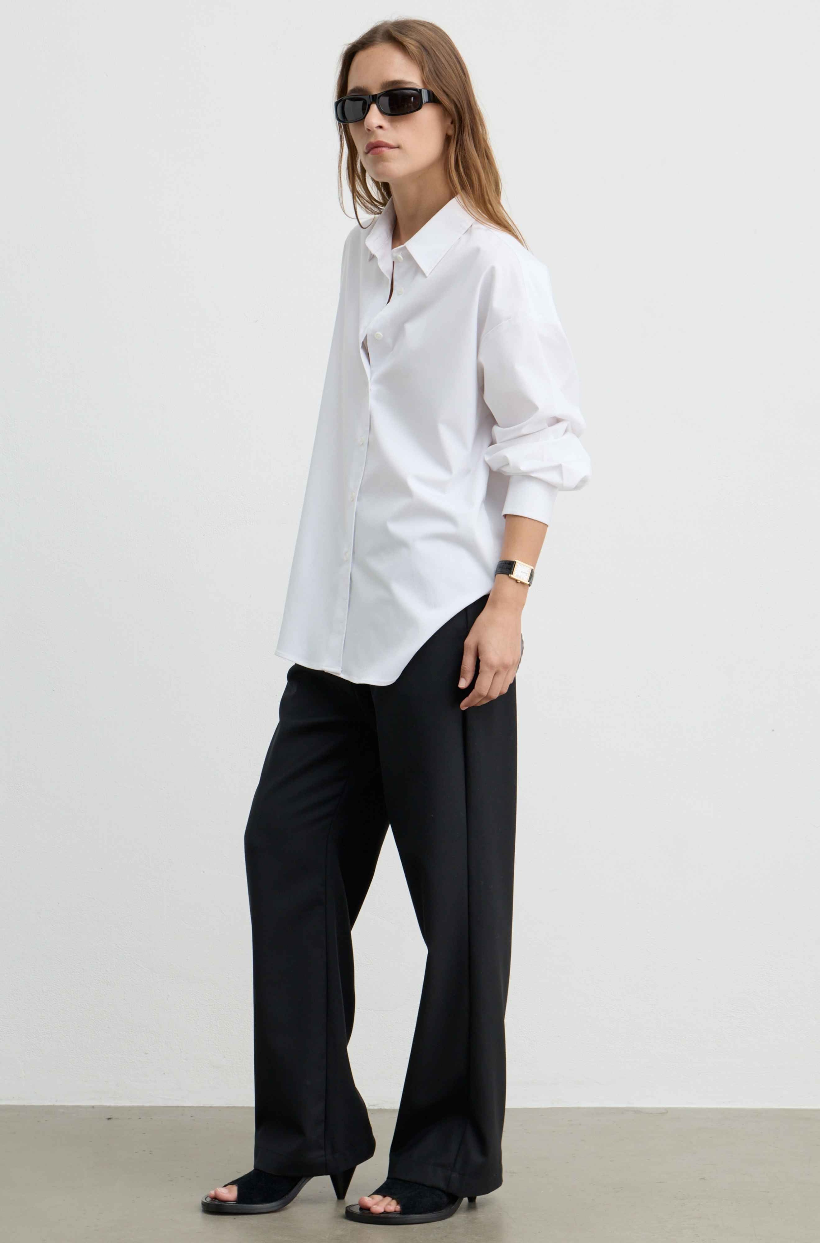 A woman modeling a white long-sleeve button-up shirt with black wide-leg trousers and black sandals, standing against a plain light gray wall.
