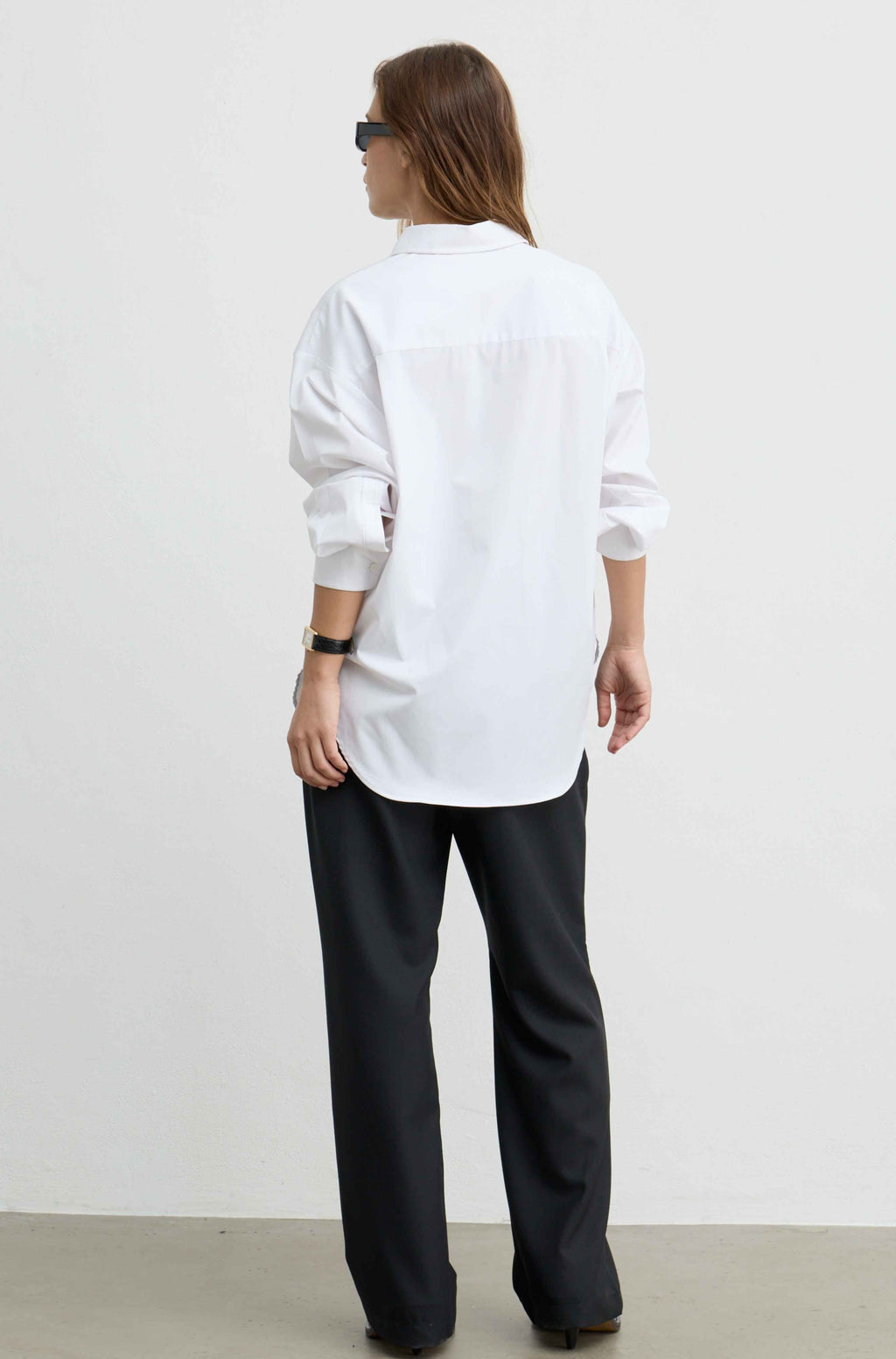 A woman shown from the back wearing a white oversized button-down shirt, black wide-leg trousers, and sunglasses against a plain white studio wall.