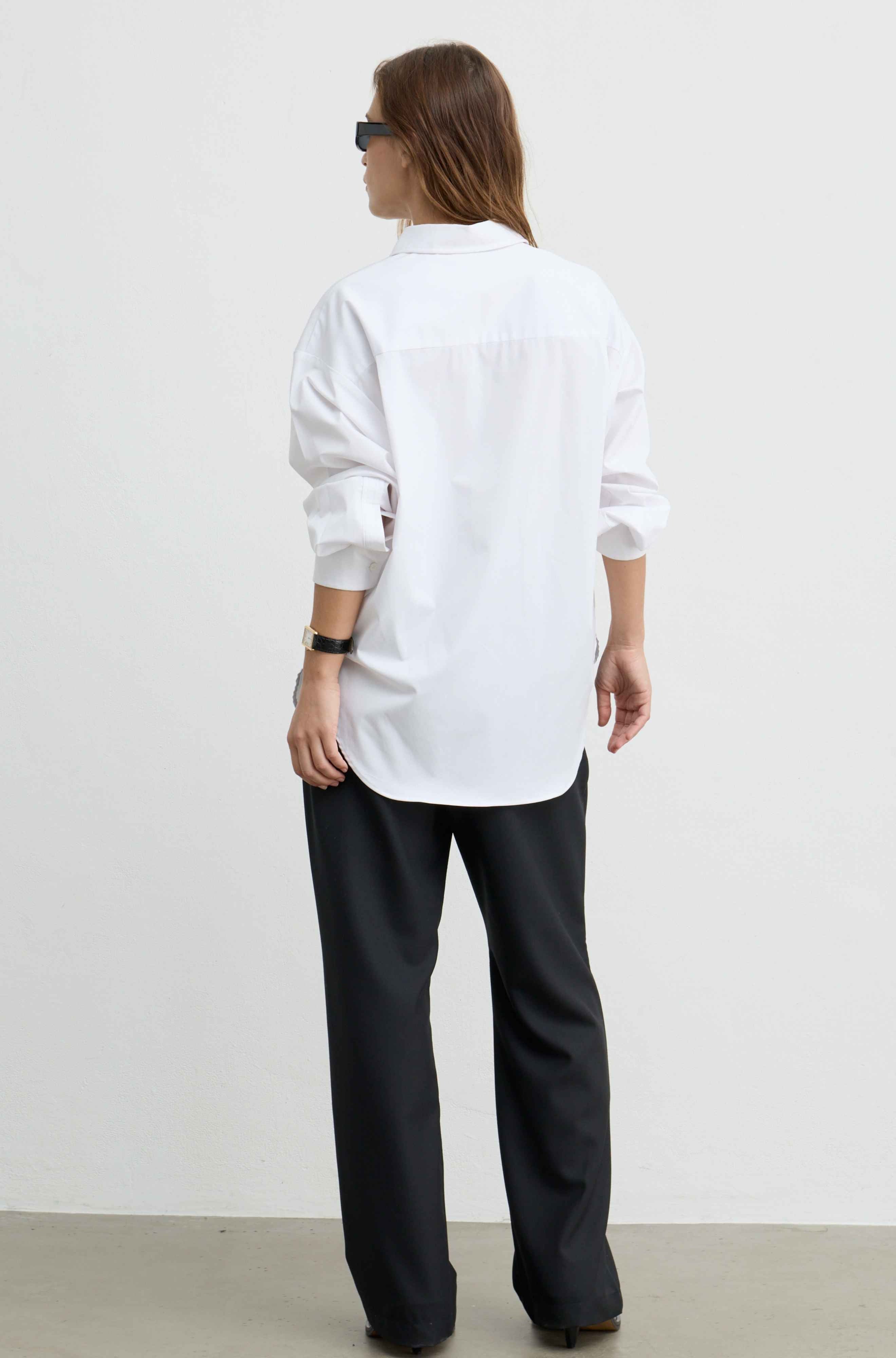 A woman shown from the back wearing a white oversized button-down shirt, black wide-leg trousers, and sunglasses against a plain white studio wall.