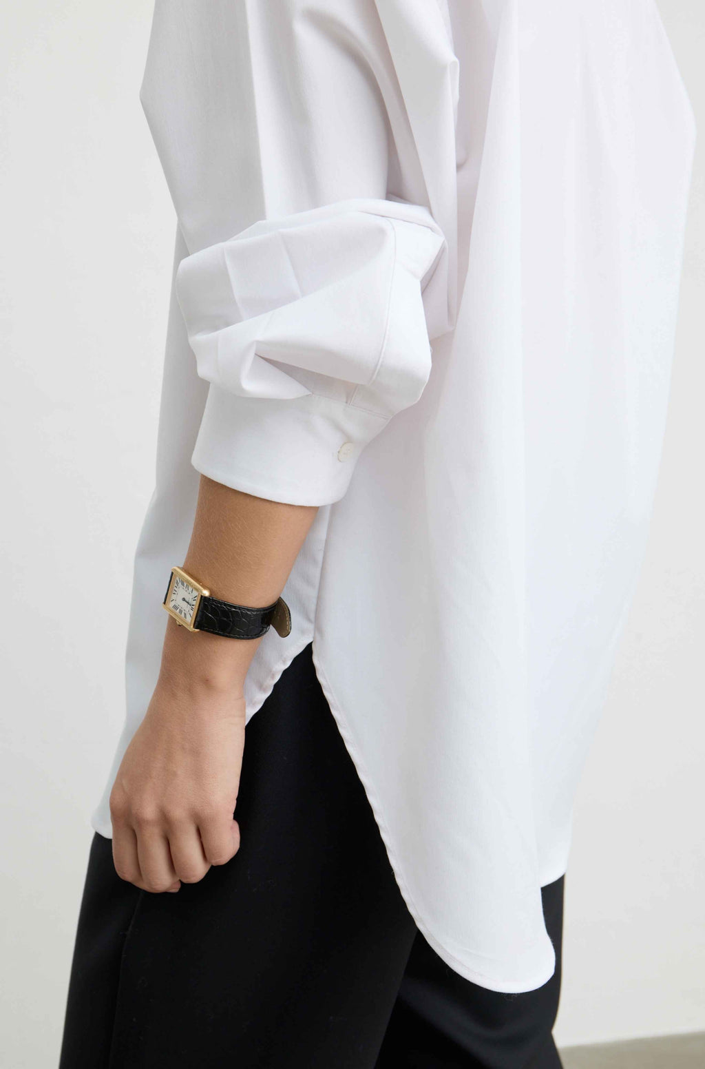 A close-up of a person wearing an oversized white long-sleeve shirt with rolled cuffs, paired with black trousers and a black leather watch with a gold-toned rectangular face.