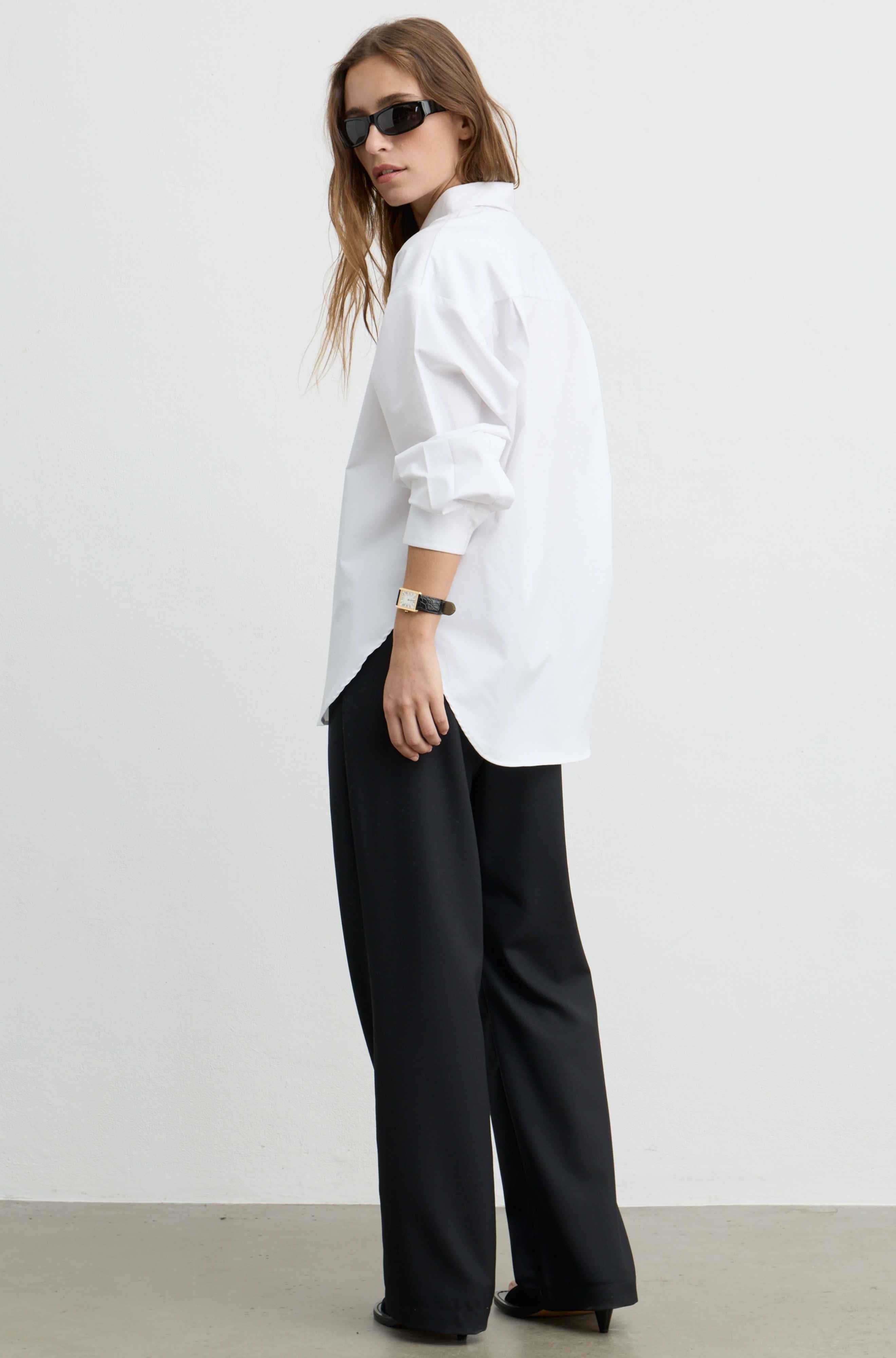 A woman modeling a white oversized long-sleeve button-up shirt paired with black wide-leg trousers, photographed against a plain light gray studio background.