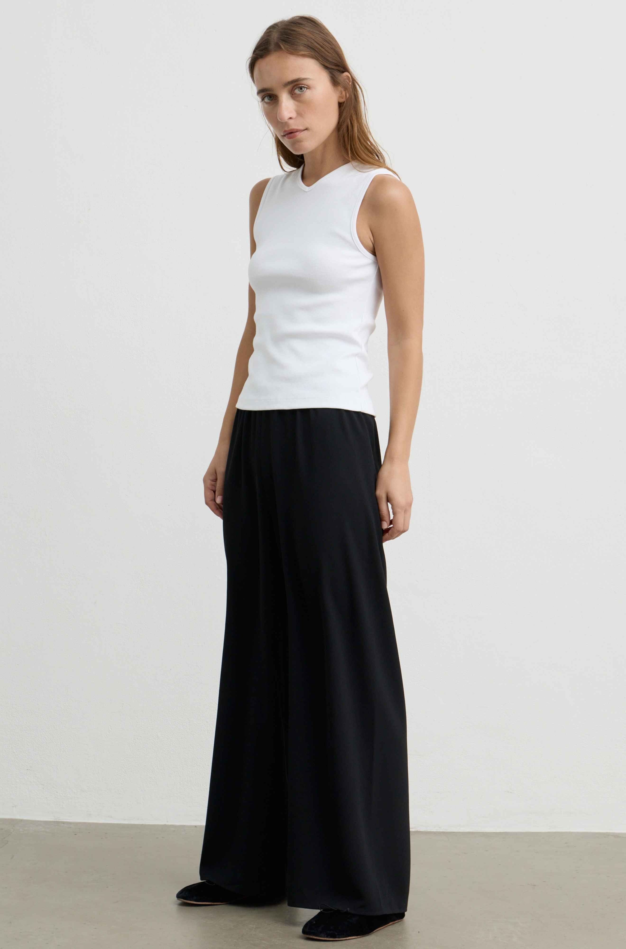 A woman modeling a sleeveless white top and wide-leg black pants against a plain light gray studio background.