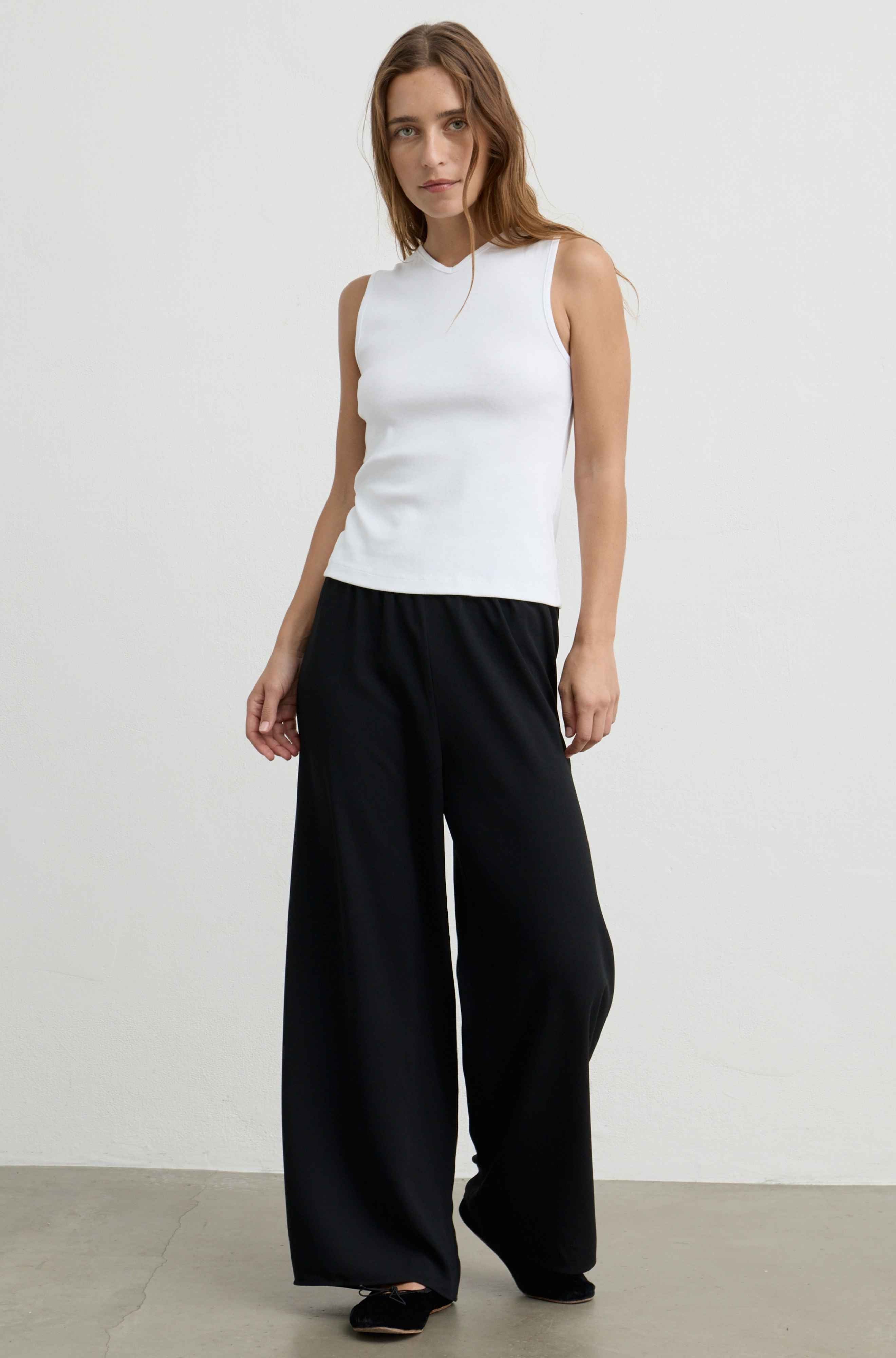 A woman modeling a sleeveless white mock-neck top paired with wide-leg black trousers, standing against a plain light gray studio background.