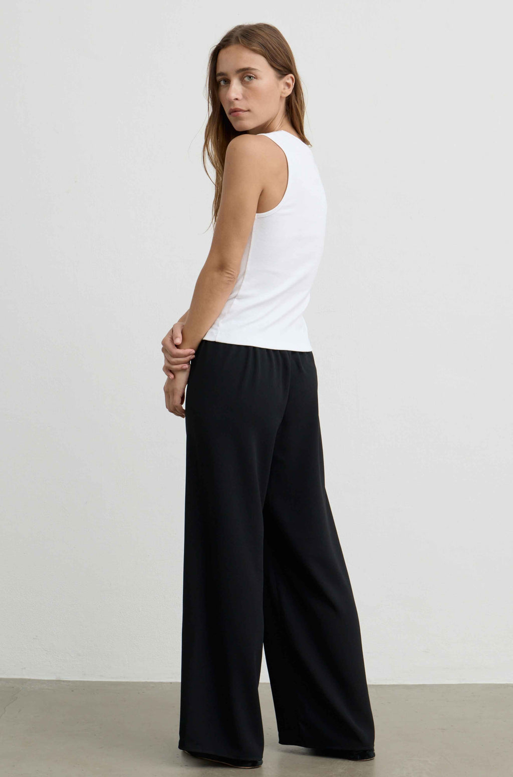 A woman wearing a white sleeveless tank top and black wide-leg pants, photographed from the back in a studio setting with a light gray wall and concrete floor.