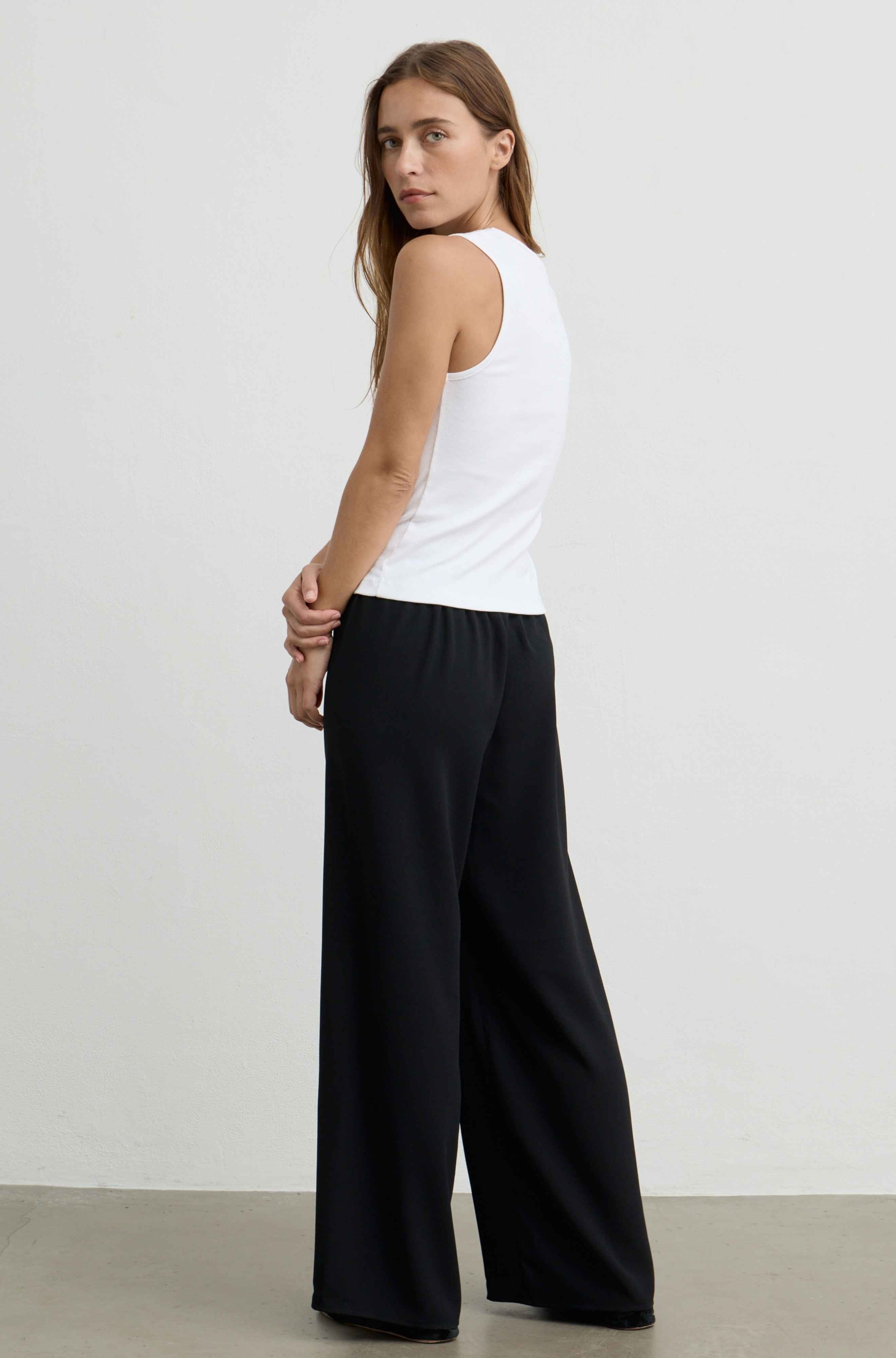 A woman wearing a white sleeveless tank top and black wide-leg pants, photographed from the back in a studio setting with a light gray wall and concrete floor.