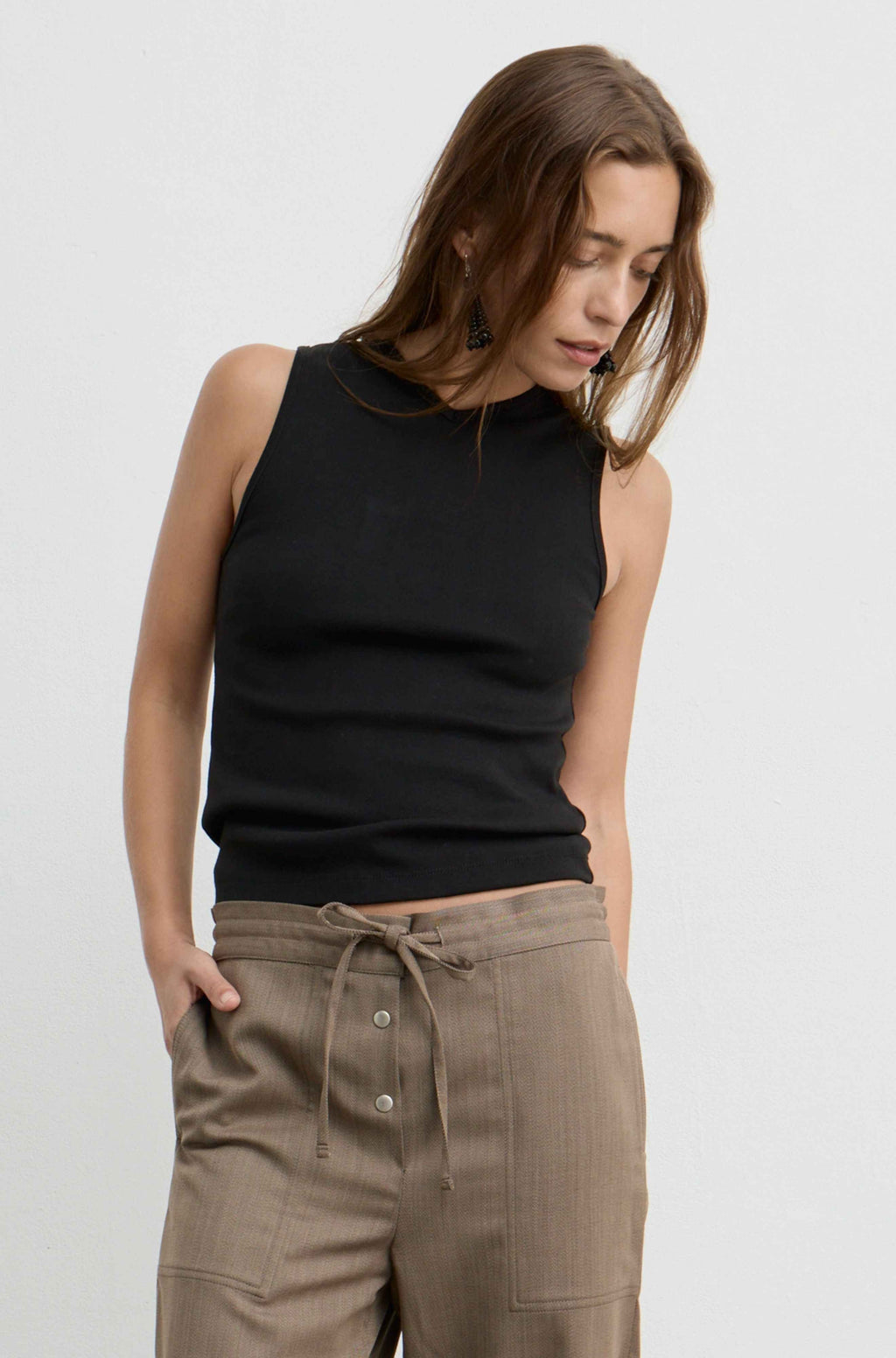A woman wearing a black sleeveless v-neck knit top and brown drawstring trousers, photographed against a clean light gray studio background.
