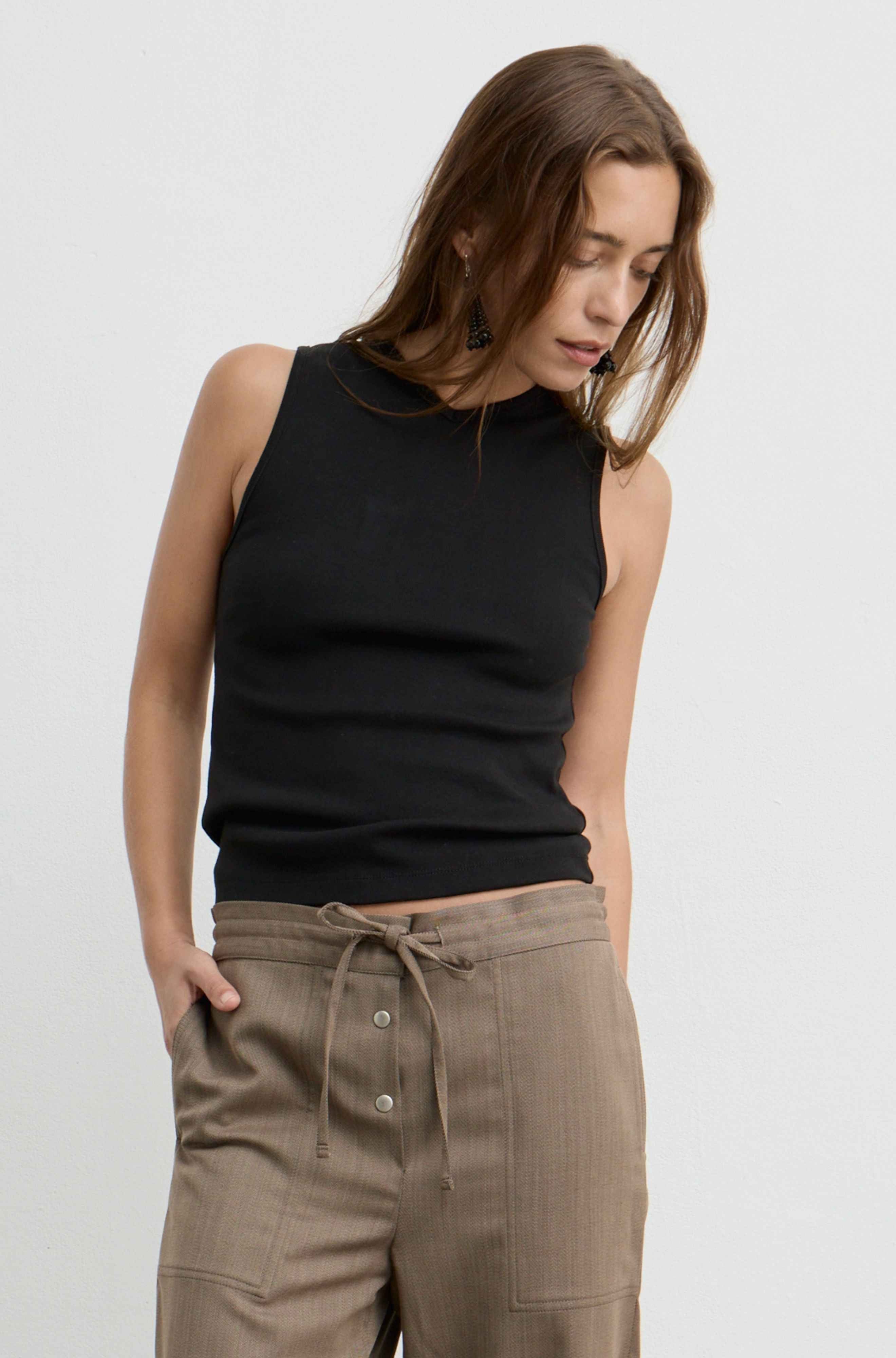 A woman wearing a black sleeveless v-neck knit top and brown drawstring trousers, photographed against a clean light gray studio background.