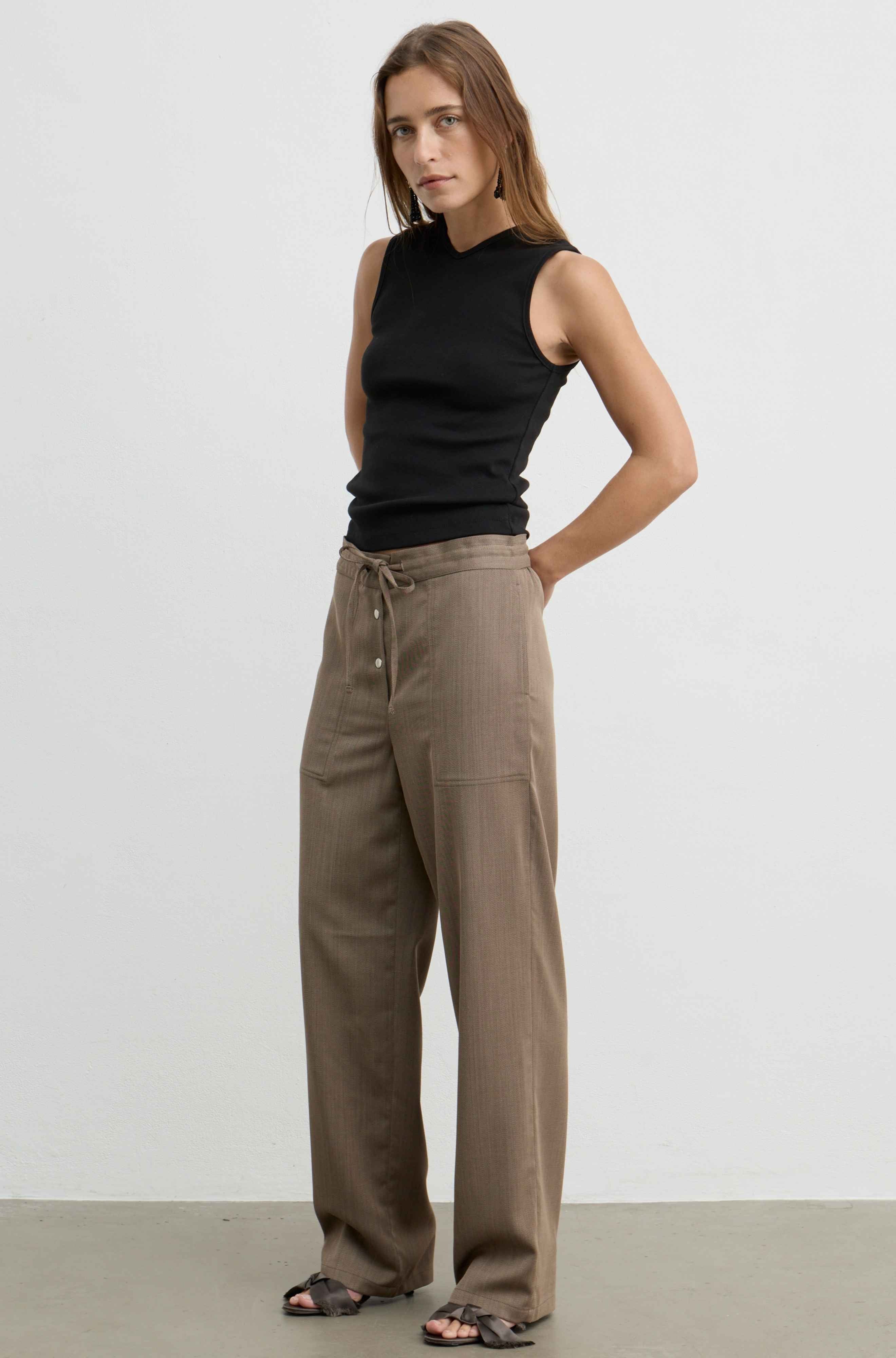 A woman modeling a black sleeveless v-neck top and brown wide-leg drawstring trousers against a plain light gray studio background.
