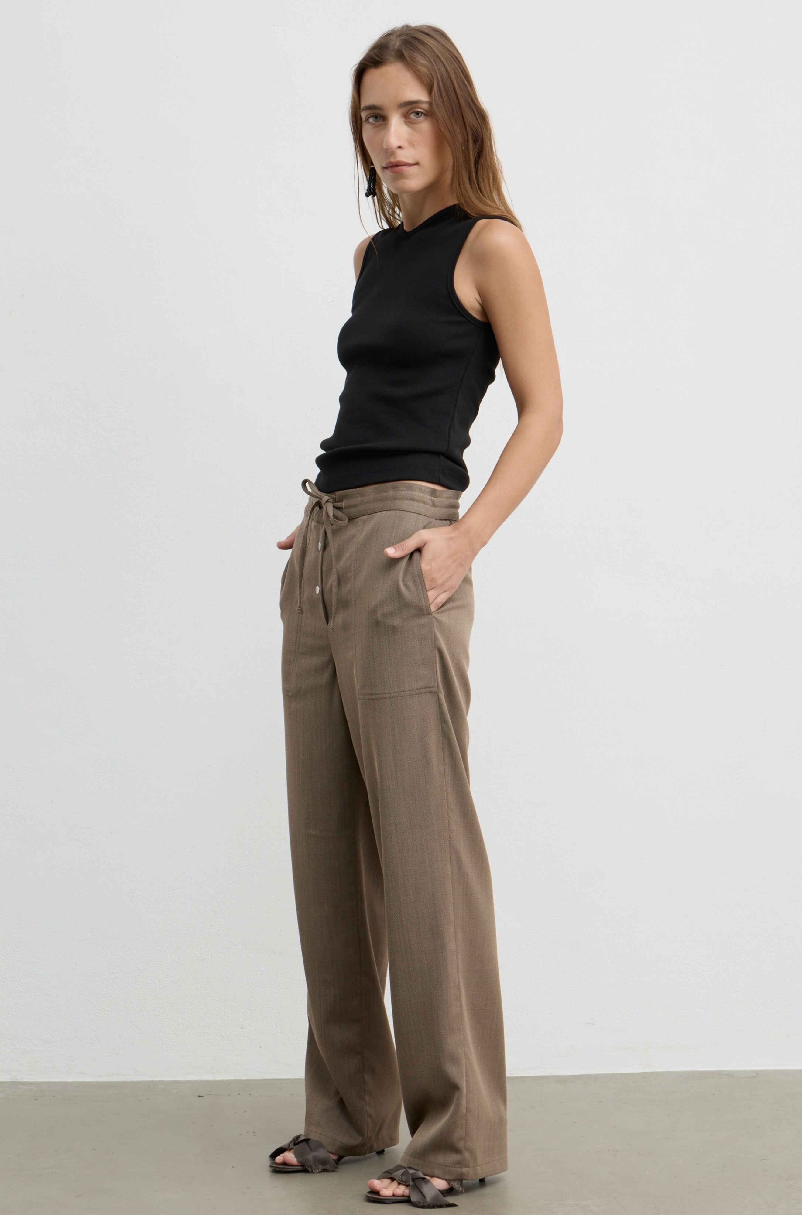 A woman modeling a sleeveless black ribbed top and wide-leg taupe pants, standing against a plain light gray studio wall.