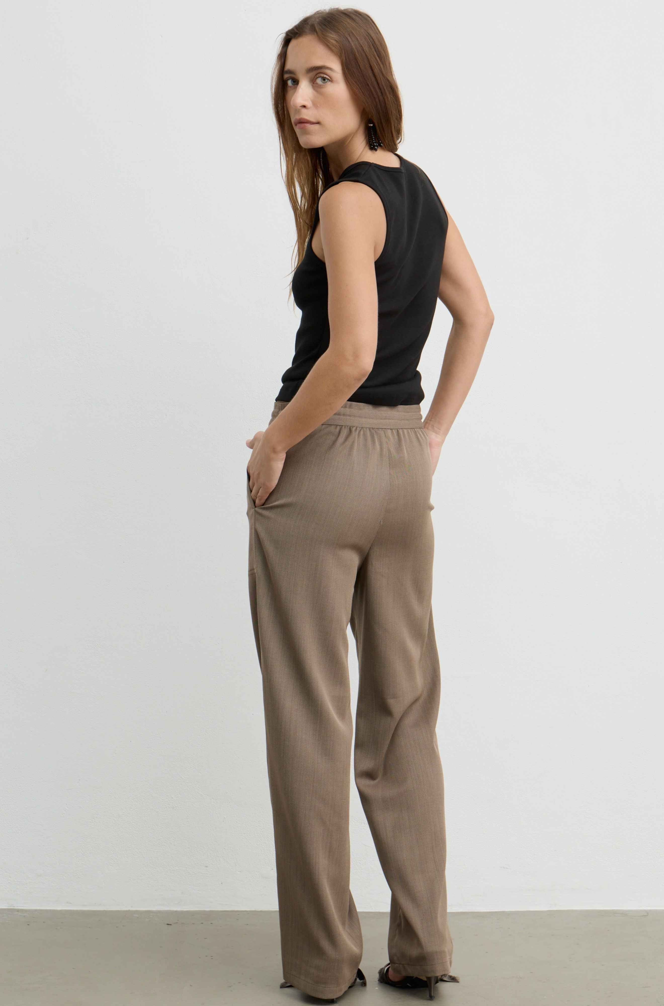 A woman modeling a pair of wide-leg taupe pants with a black sleeveless top, photographed from the back against a plain light gray studio background.