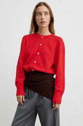 A woman modeling a bright red long-sleeve knit cardigan with white buttons, styled with a dark brown wrap skirt and gray trousers, against a plain light gray studio background.