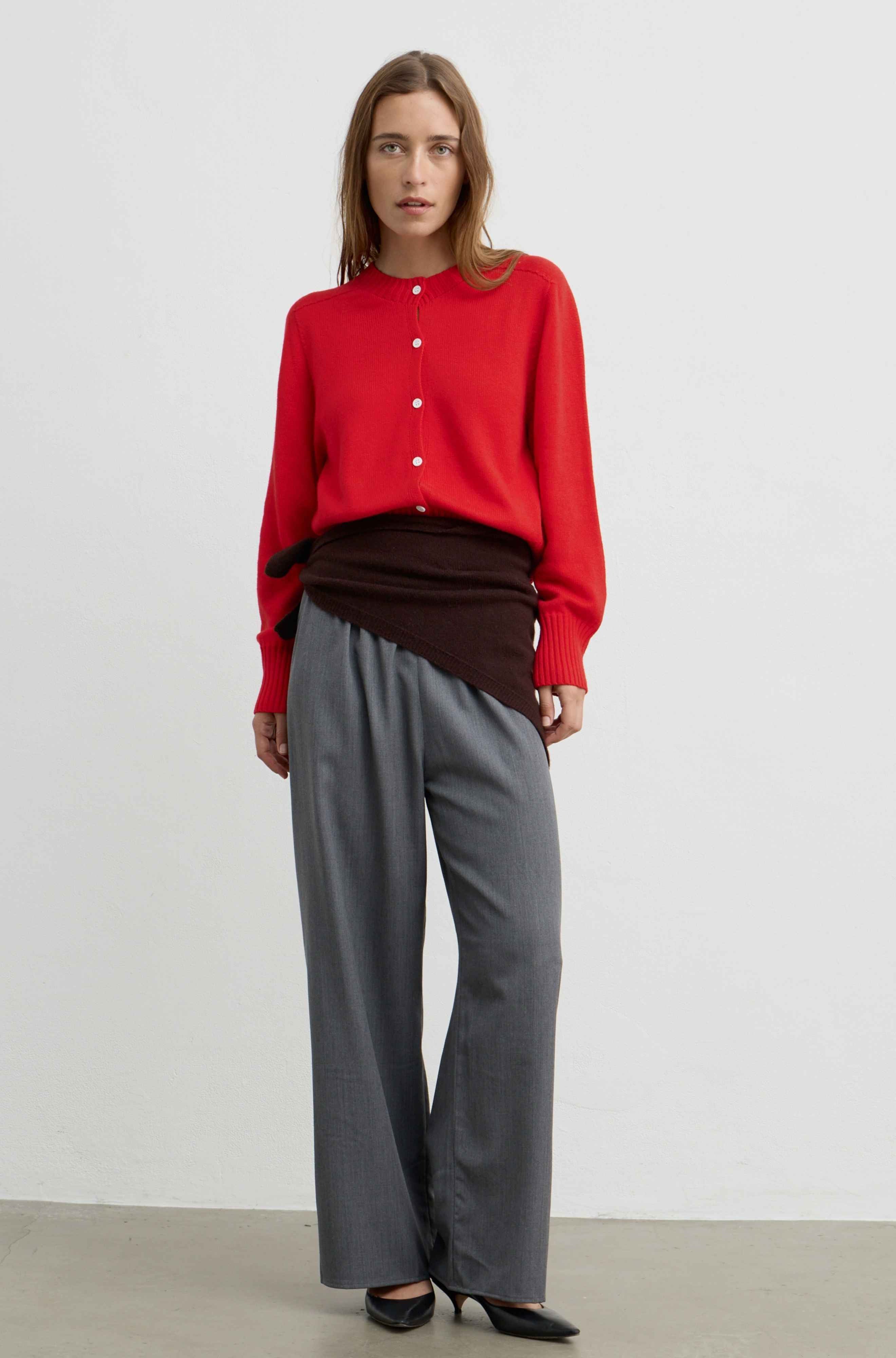 A woman modeling a red long-sleeve button-front top with a dark brown draped layer, paired with gray wide-leg trousers and black pointed heels, against a plain light gray studio background.