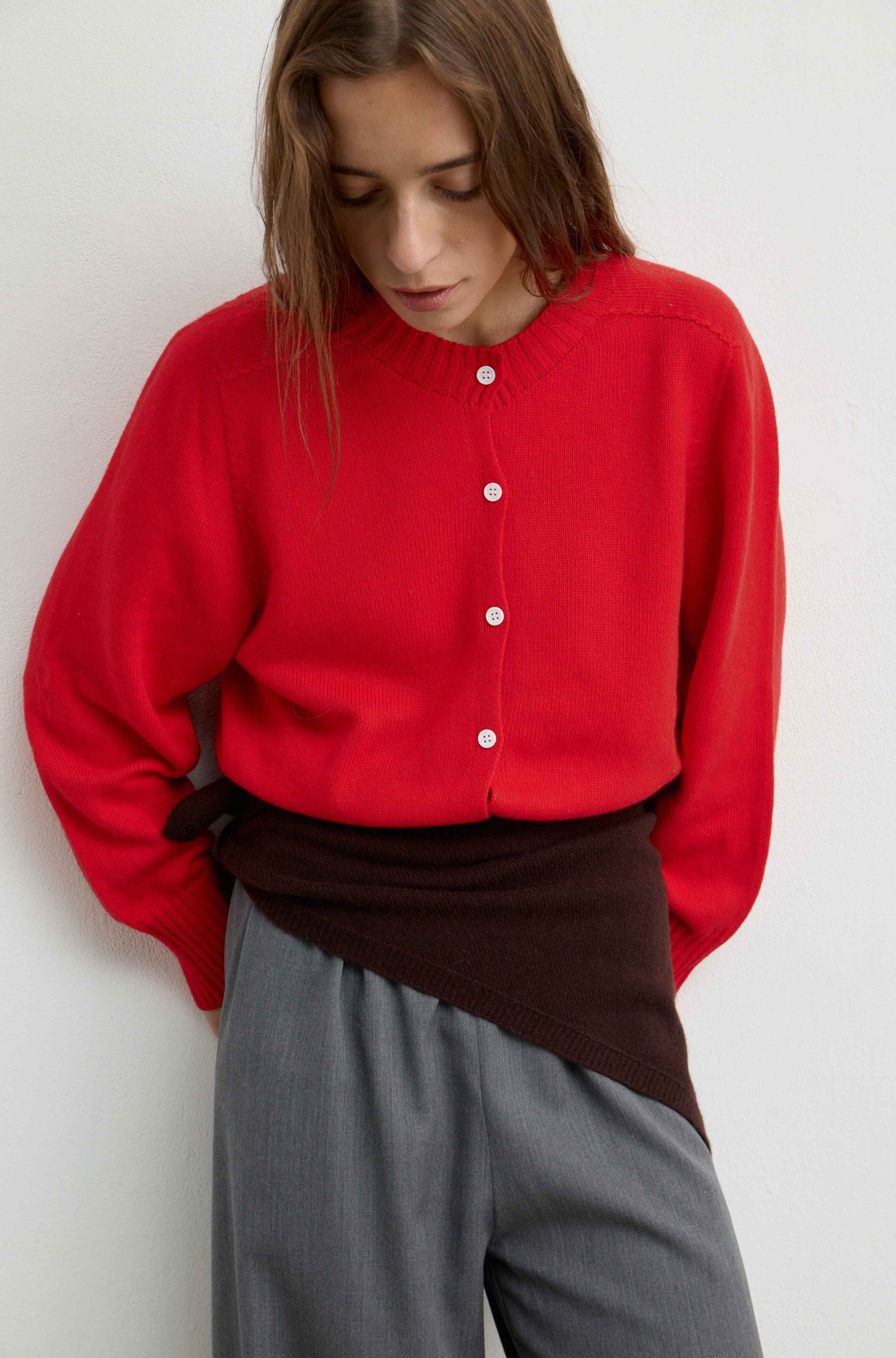 A woman wearing a bright red button-front knit cardigan with white buttons, layered over a dark brown wrap-style sweater and paired with gray trousers, photographed against a plain light background.