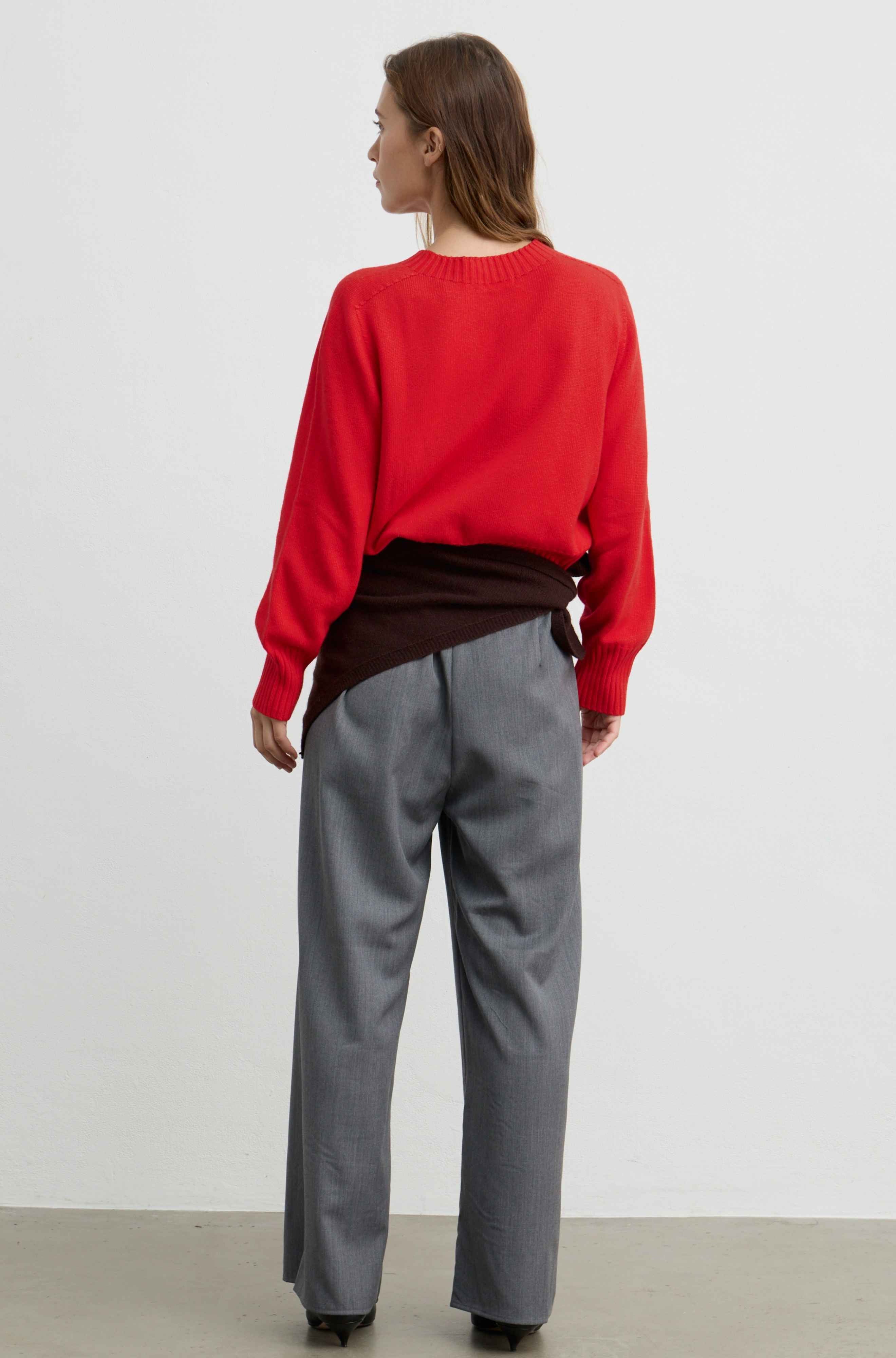 A woman is shown from behind wearing a bright red knit sweater, a dark brown shirt tucked in, and gray wide-leg trousers against a plain white studio background.