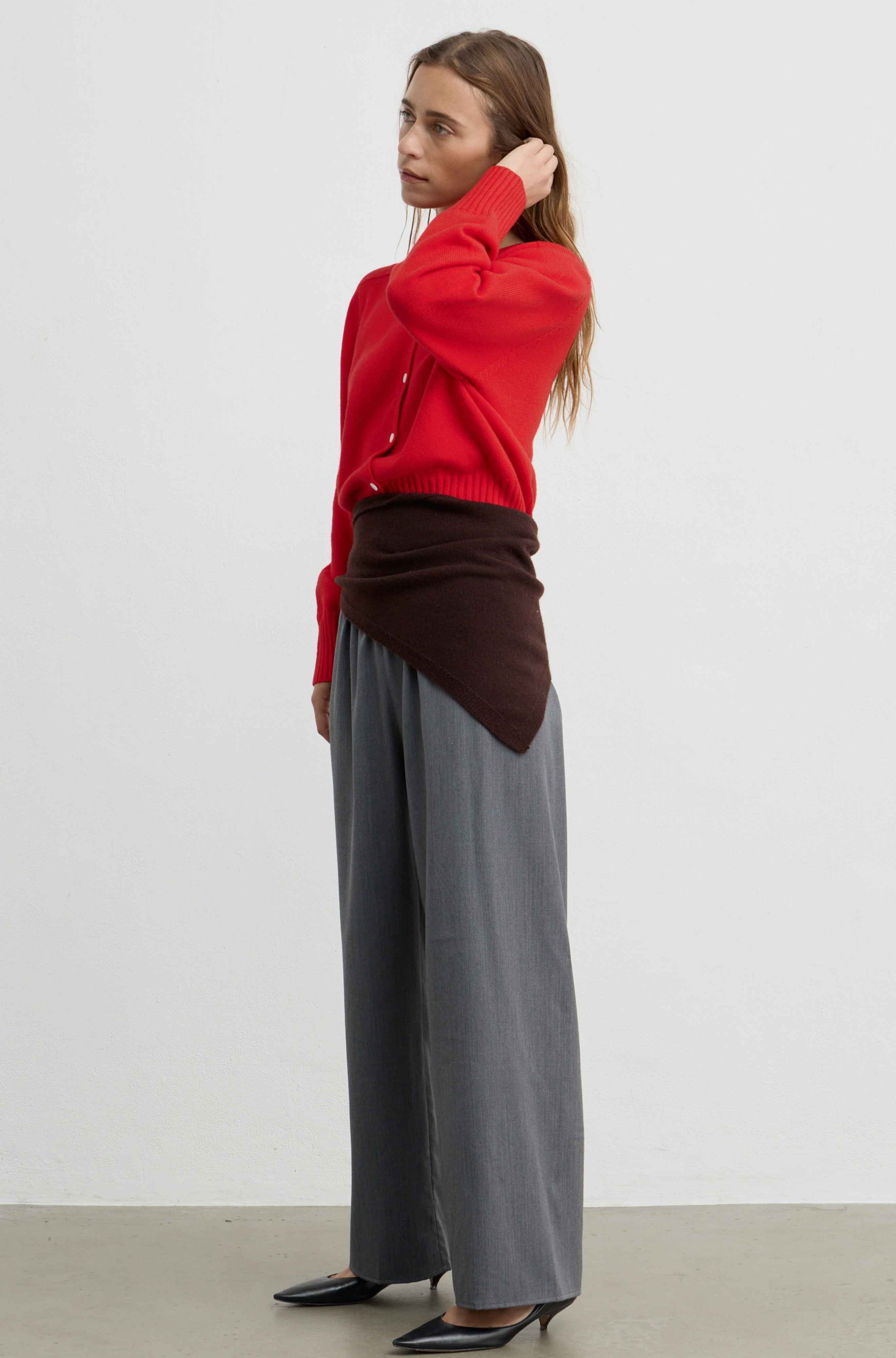 A woman stands in profile wearing a bright red knit cardigan, a dark brown wrap draped around her waist, wide-leg gray trousers, and black pointed heels against a plain light gray studio background.