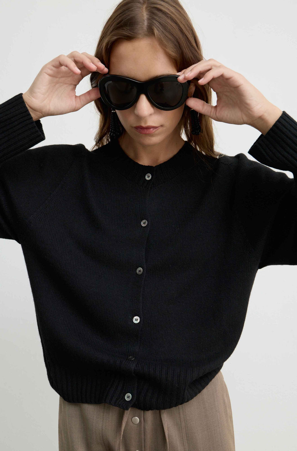 A woman wearing a black ribbed button-up cardigan sweater and beige trousers, holding black sunglasses in front of her eyes against a plain light gray background.