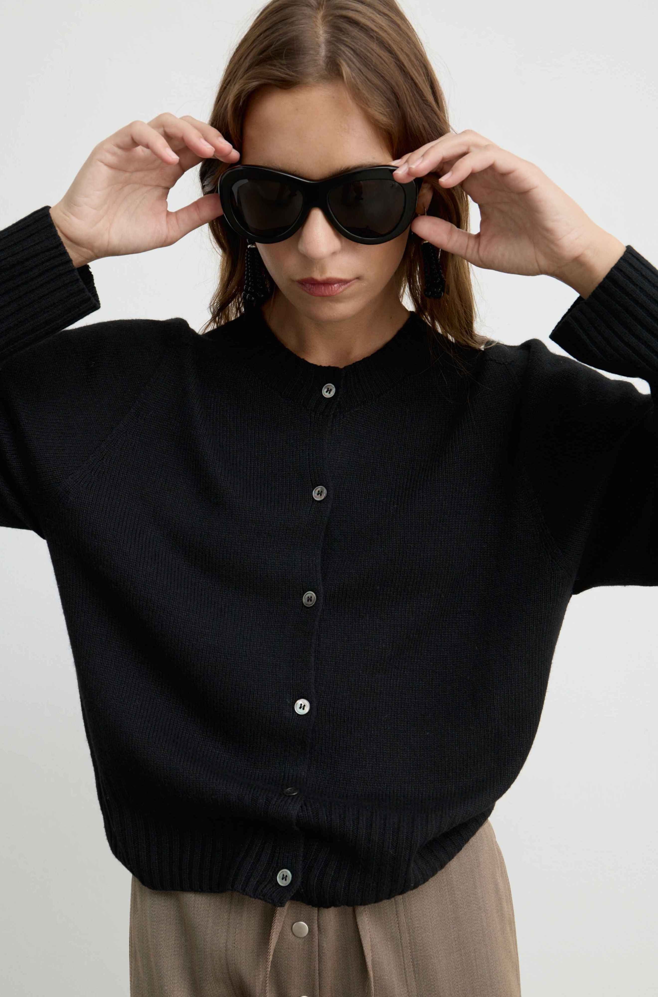 A woman wearing a black ribbed button-up cardigan sweater and beige trousers, holding black sunglasses in front of her eyes against a plain light gray background.