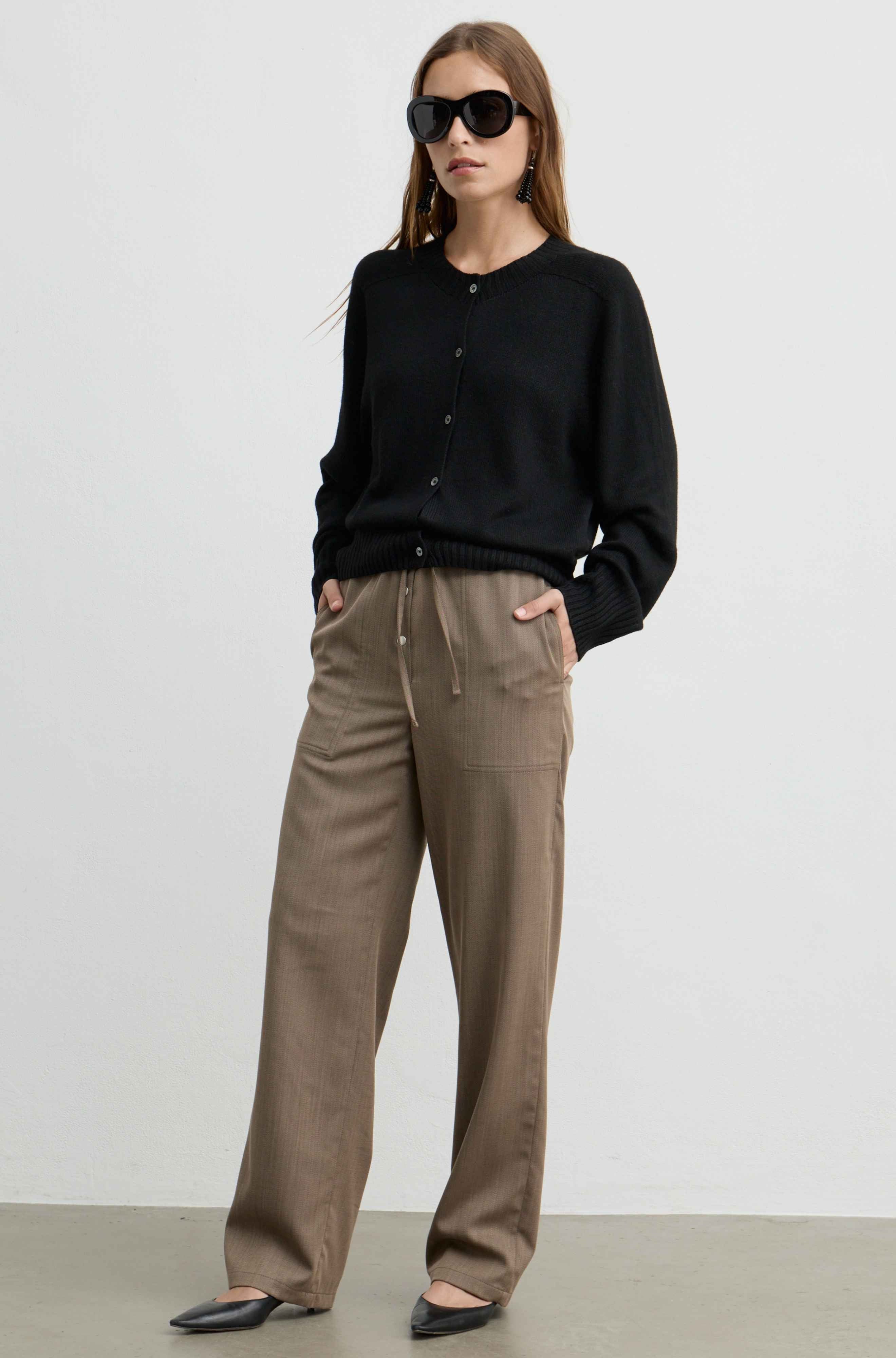A woman modeling a black knit cardigan and wide-leg taupe trousers with black flats, standing against a plain light gray studio background.