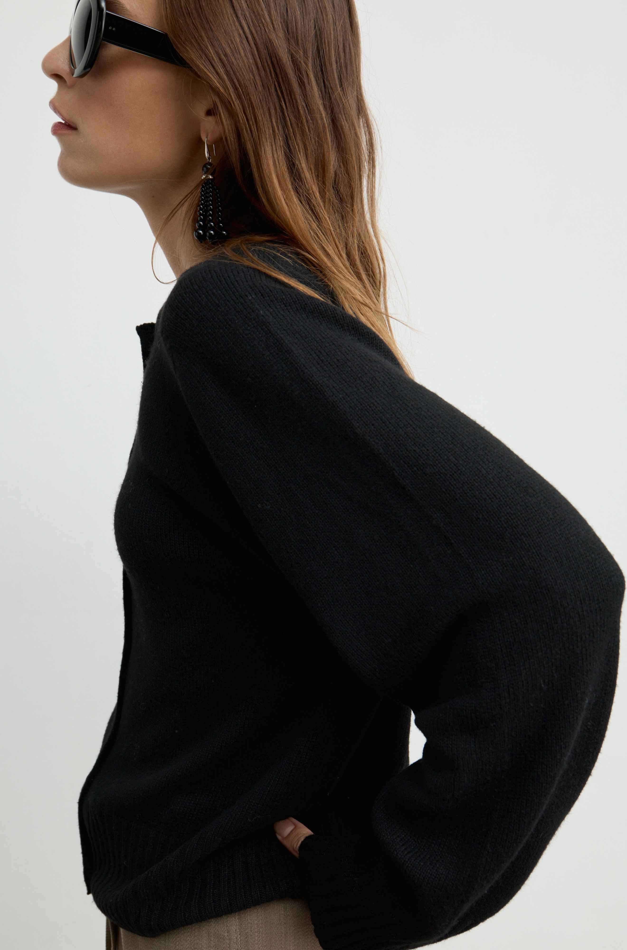 A woman shown in side profile wearing a black knit sweater, black sunglasses, and dangling black earrings against a plain light background.