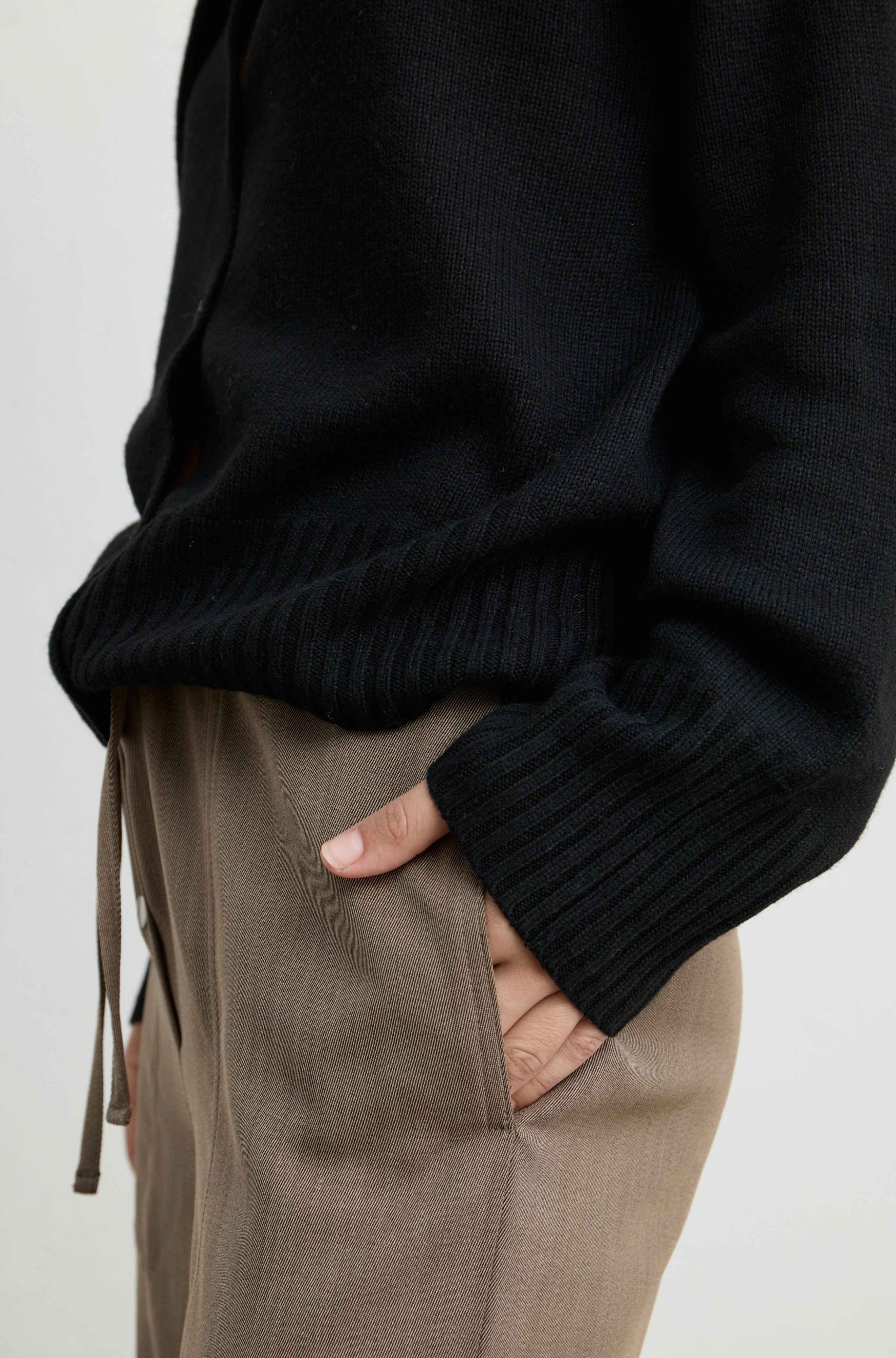 Close-up of a person wearing a black ribbed knit sweater and taupe drawstring trousers, with one hand in a pocket against a plain light background.