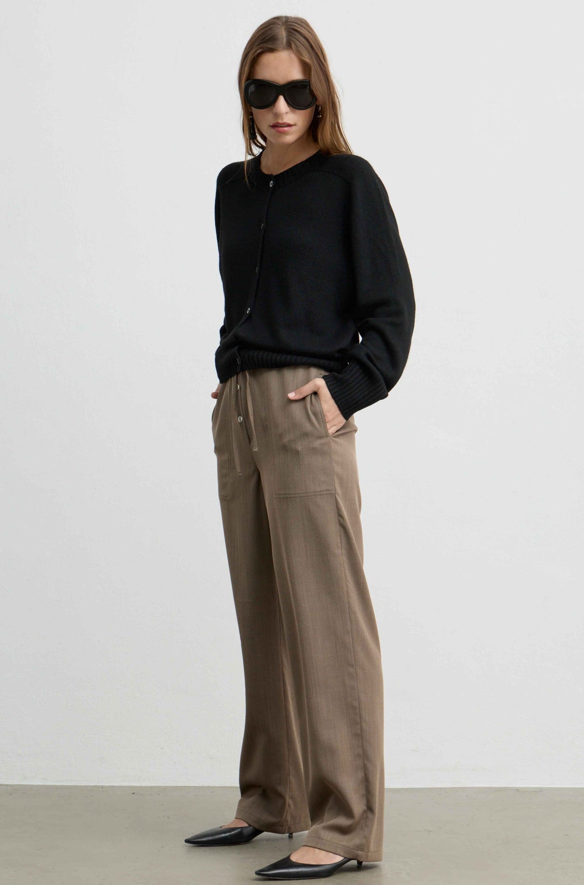 A woman modeling a black long-sleeve knit top and wide-leg taupe trousers, styled with black pointed flats against a plain light gray studio background.