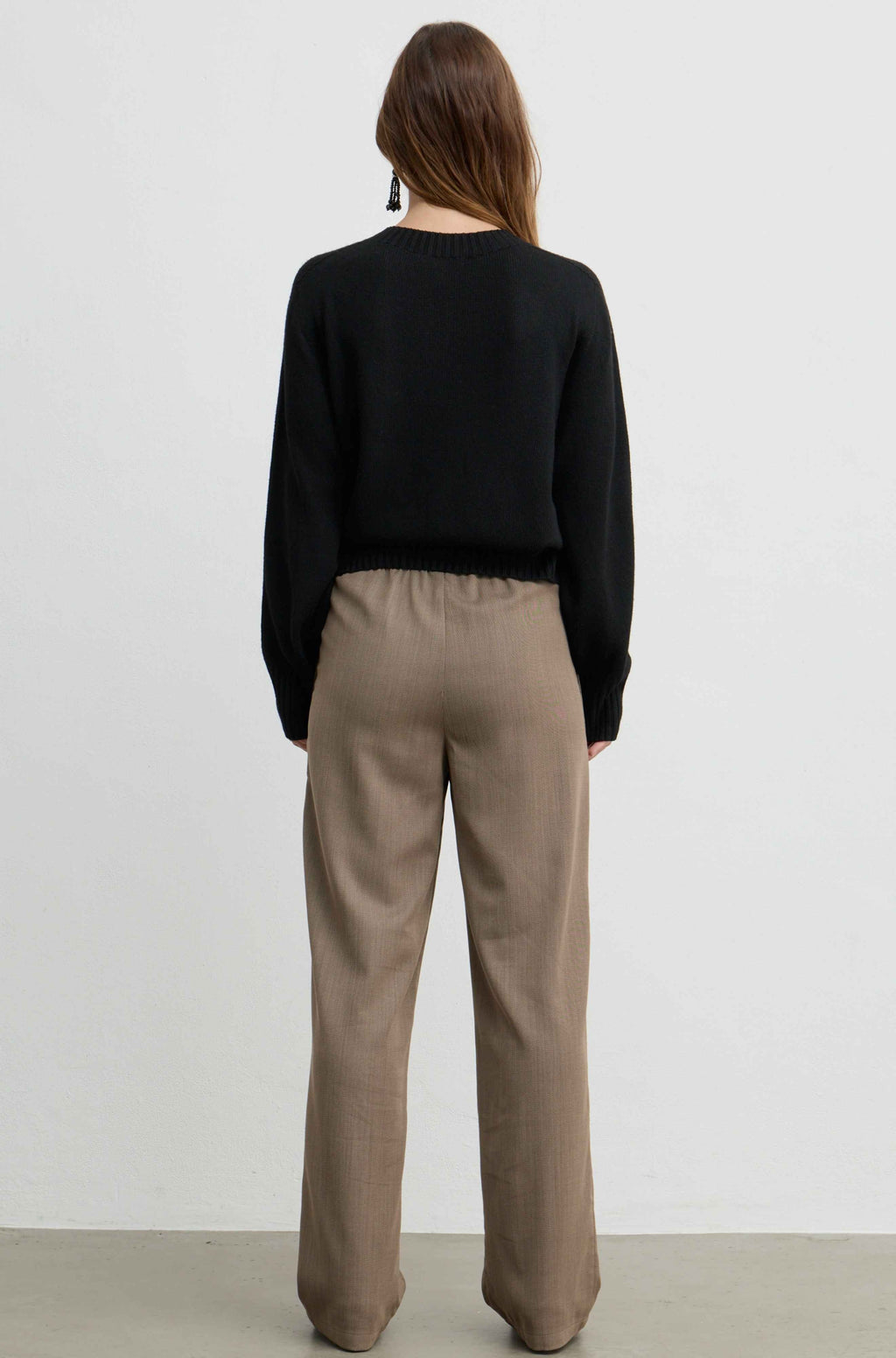 A woman viewed from behind wearing a black long-sleeve knit top and beige wide-leg trousers, standing against a plain light gray wall.
