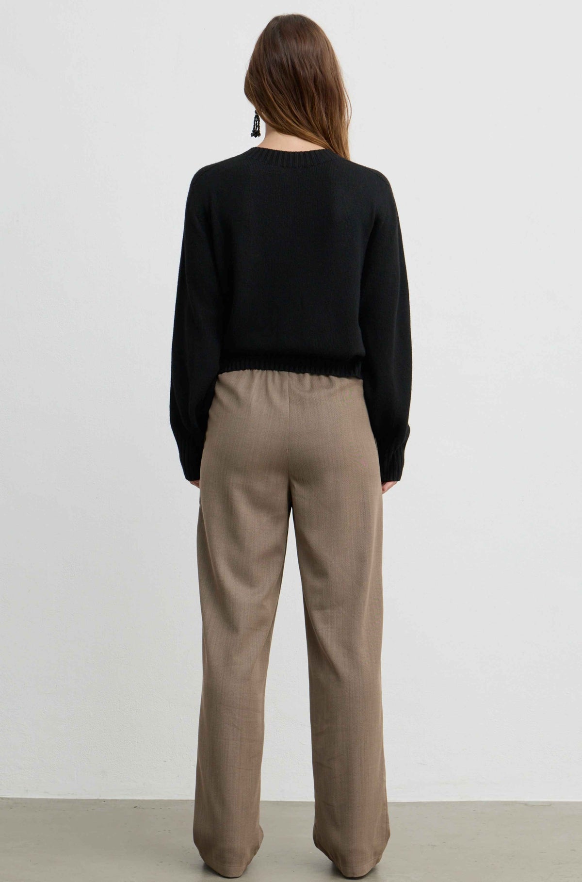 A woman viewed from behind wearing a black long-sleeve knit top and beige wide-leg trousers, standing against a plain light gray wall.
