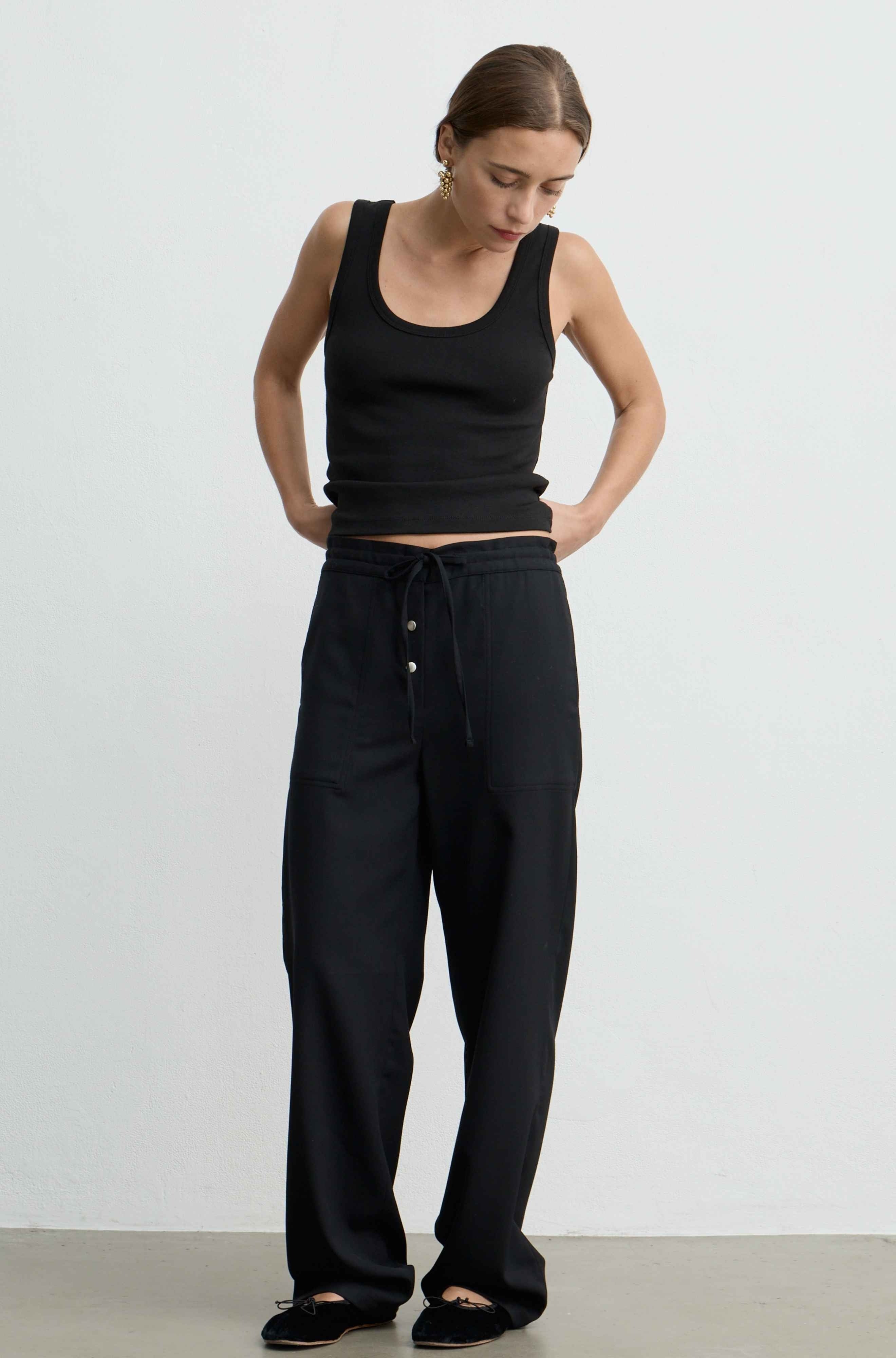 A woman modeling a black ribbed tank top and black wide-leg drawstring pants against a plain light gray studio background.