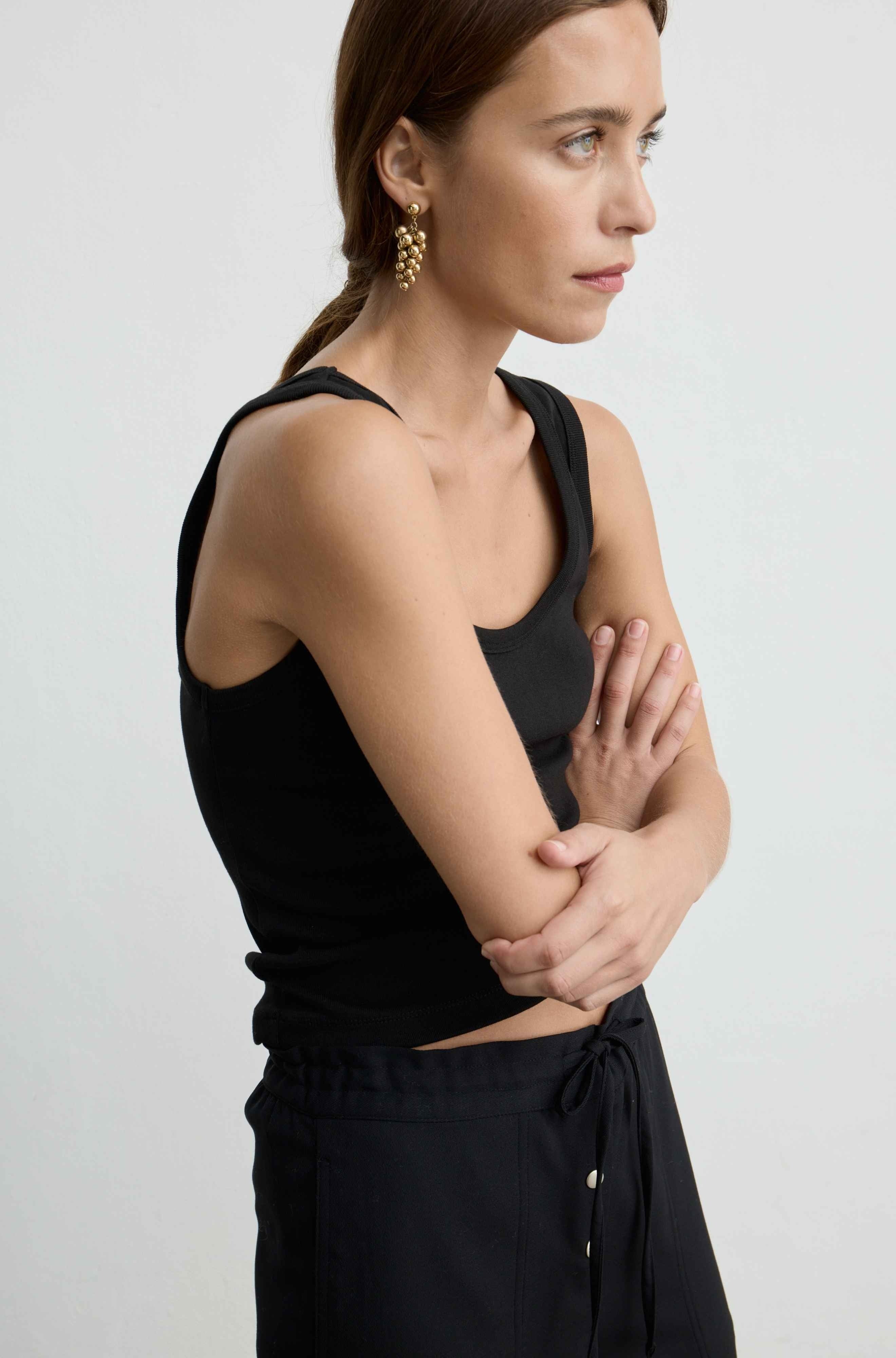 A woman wearing a black sleeveless tank and black drawstring pants, shown in profile against a light gray studio background.