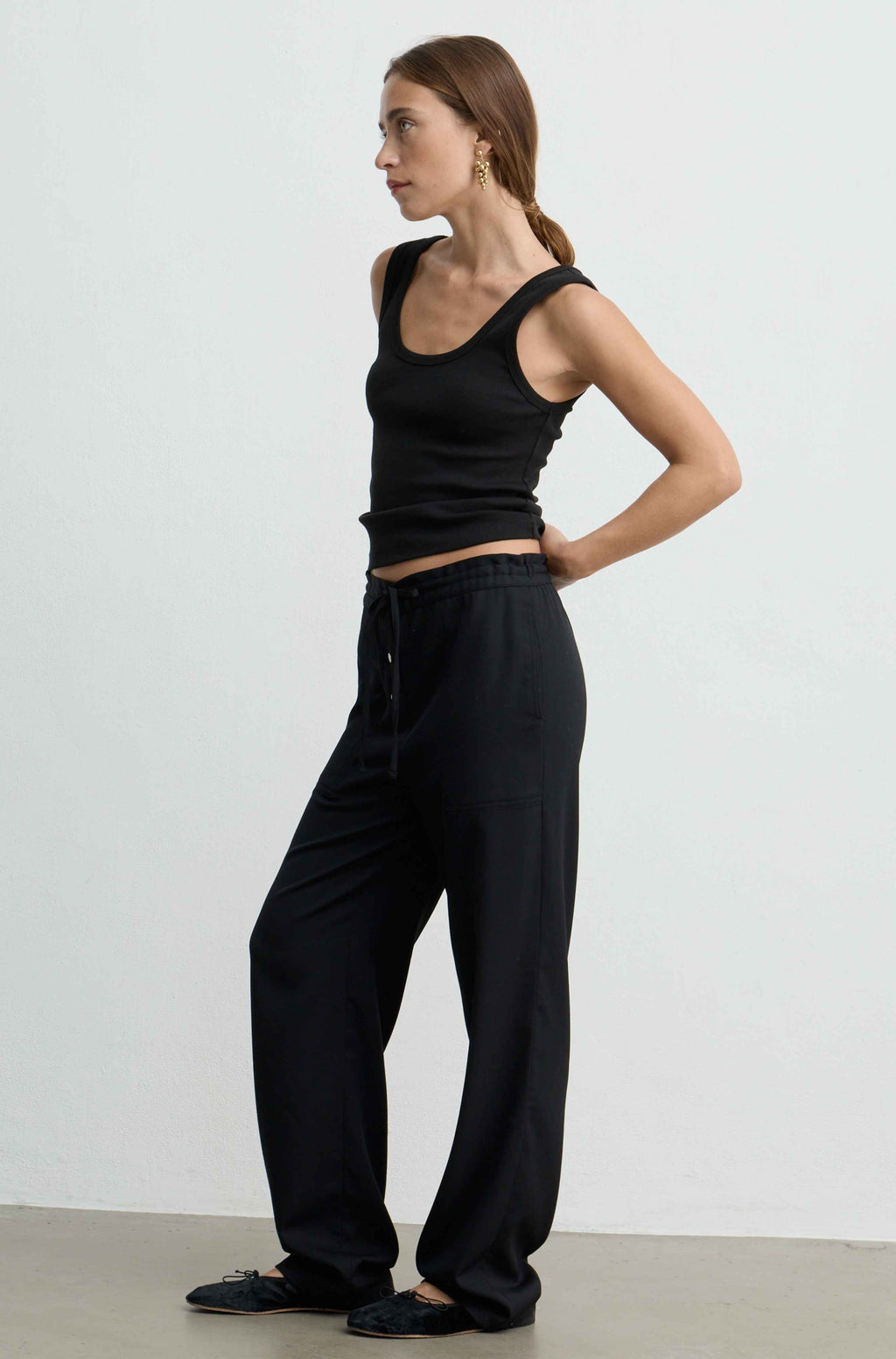 A woman modeling a black ribbed tank top and matching black wide-leg drawstring pants against a plain light gray studio background.