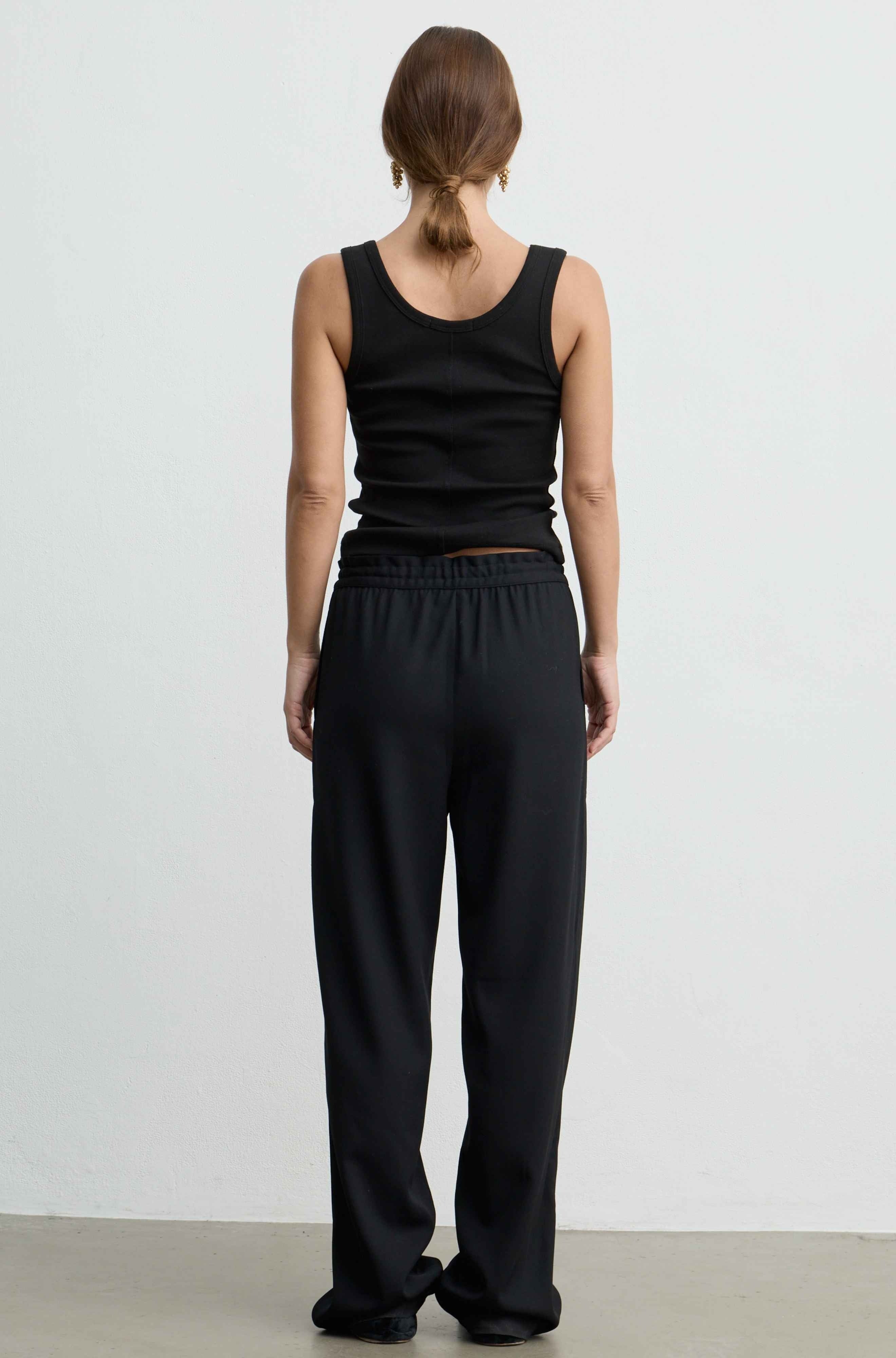 A woman viewed from the back wearing a black sleeveless tank top and loose black wide-leg pants against a plain light gray studio background.