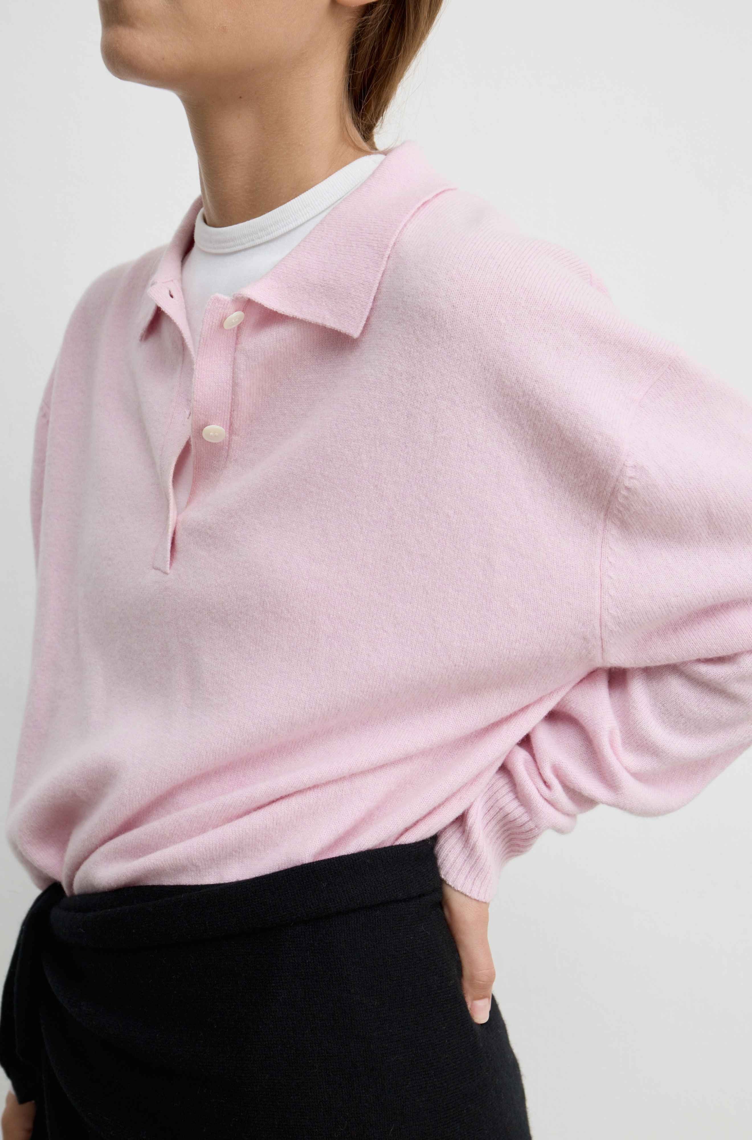 A close-up of a model wearing a light pink knit polo sweater over a white crewneck t-shirt and black wrap scarf tied around her waist, set against a plain light gray background.