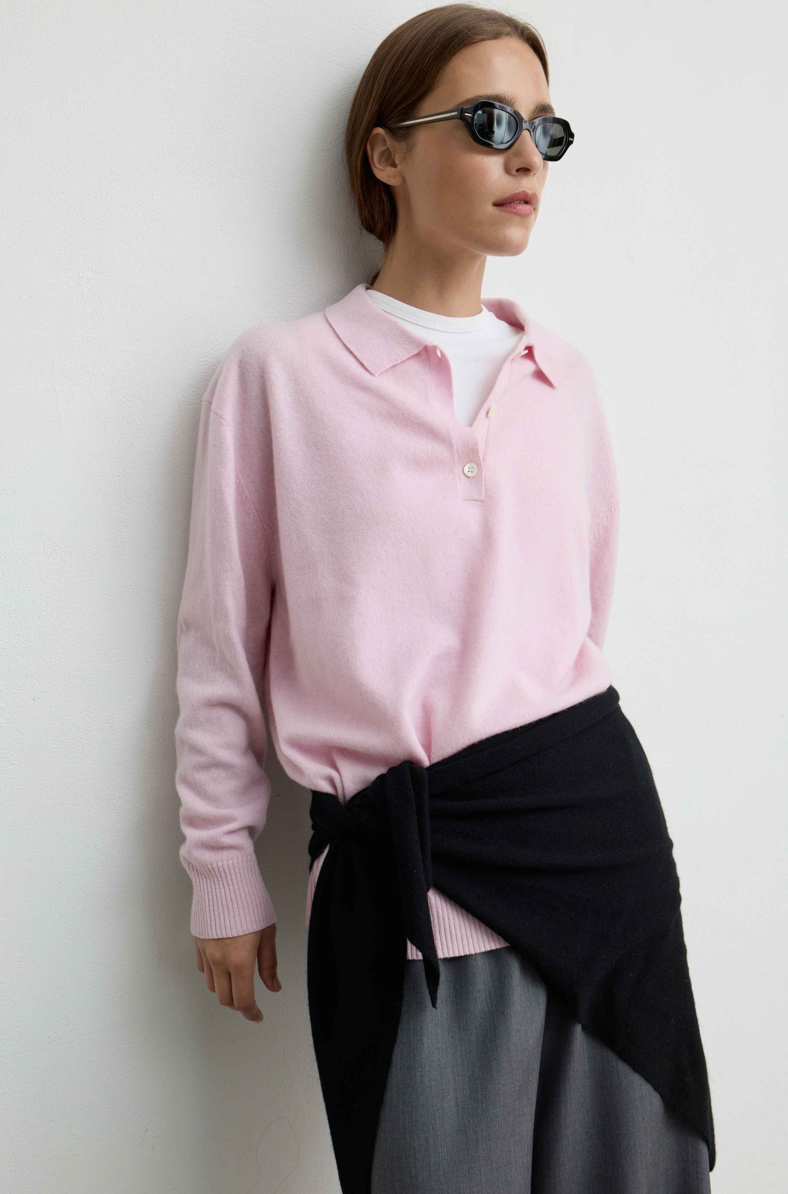 A woman wearing a light pink knit polo sweater over a white t-shirt, styled with a black wrap scarf and gray trousers, leaning against a white wall.