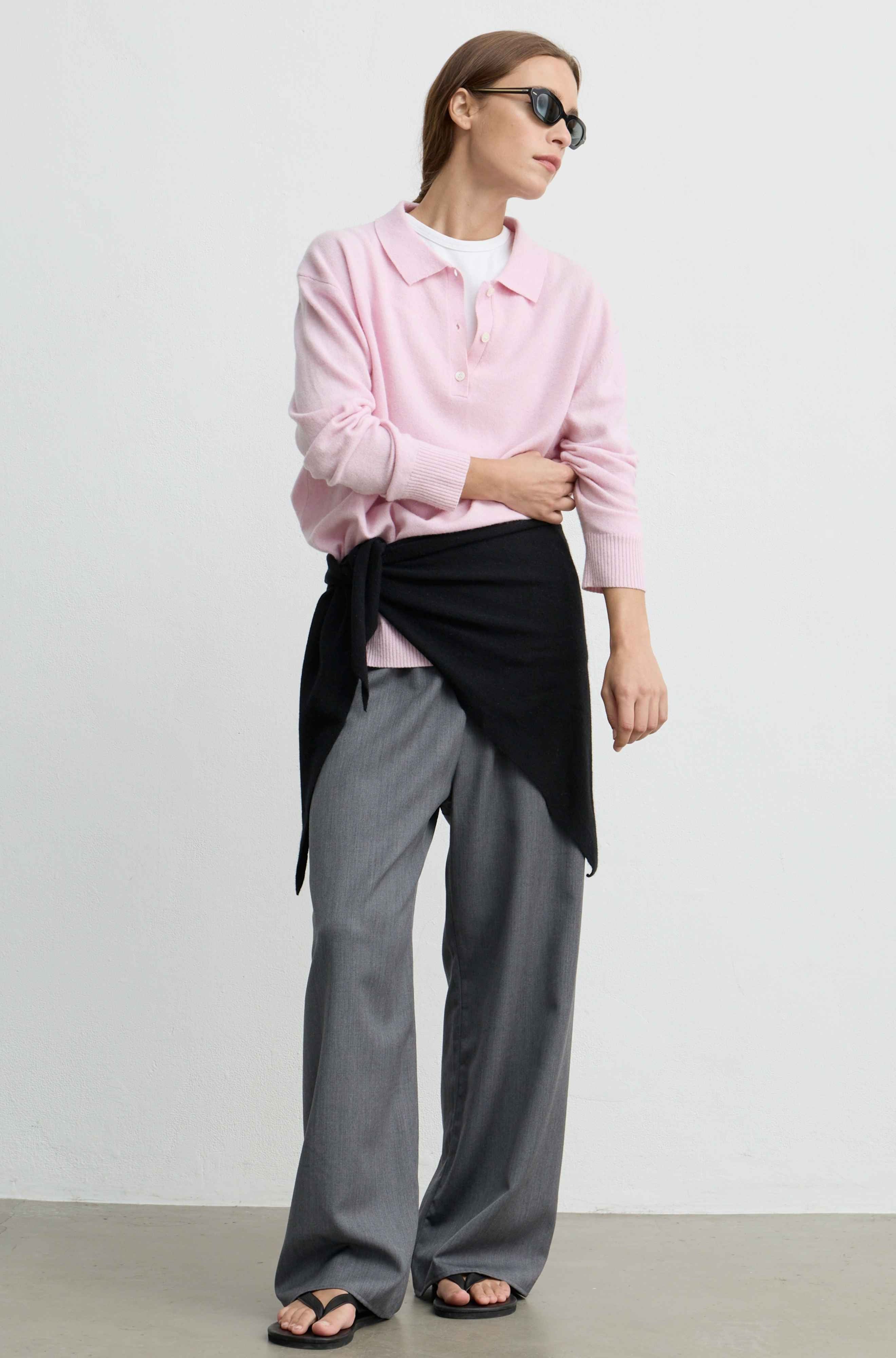 A woman modeling a light pink long-sleeve polo shirt, styled with a black wrap scarf tied at the waist, gray wide-leg trousers, and black sandals against a plain light gray studio background.
