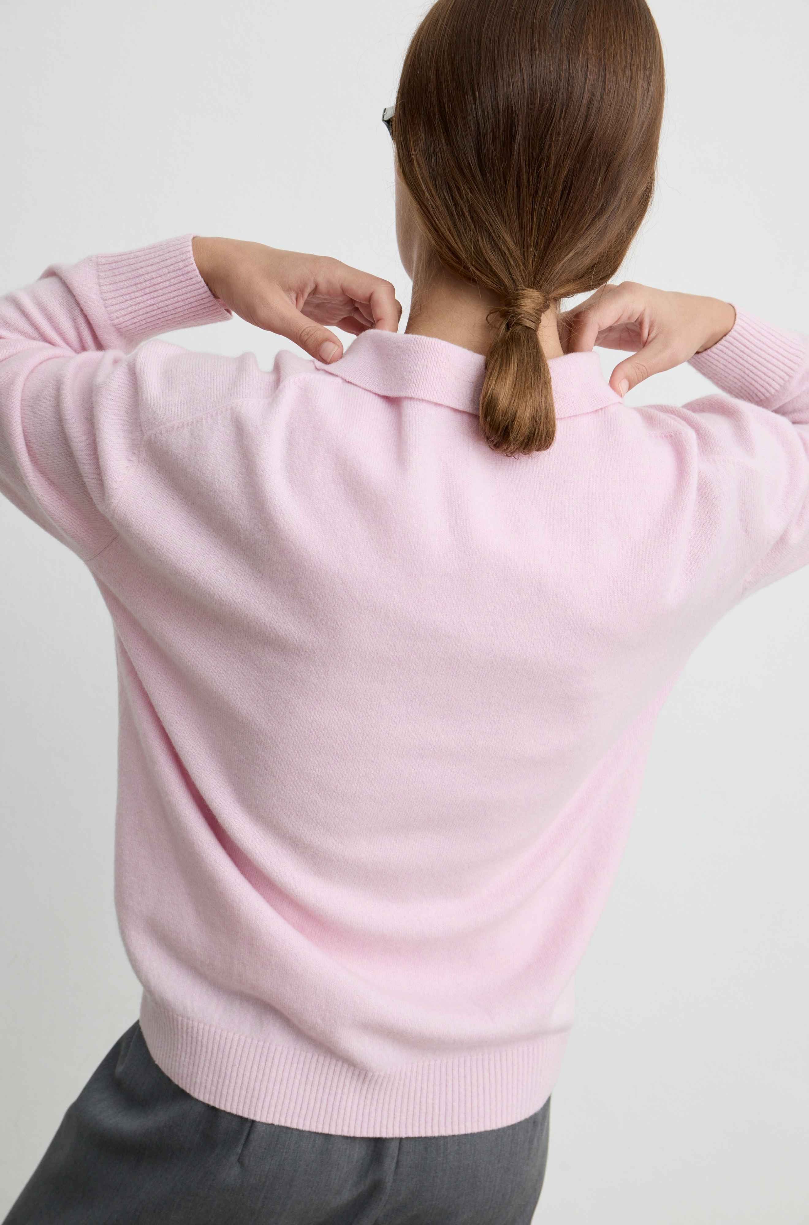 A woman viewed from behind wearing a light pink knit sweater and dark gray trousers, adjusting the collar of the sweater against a plain light gray background.