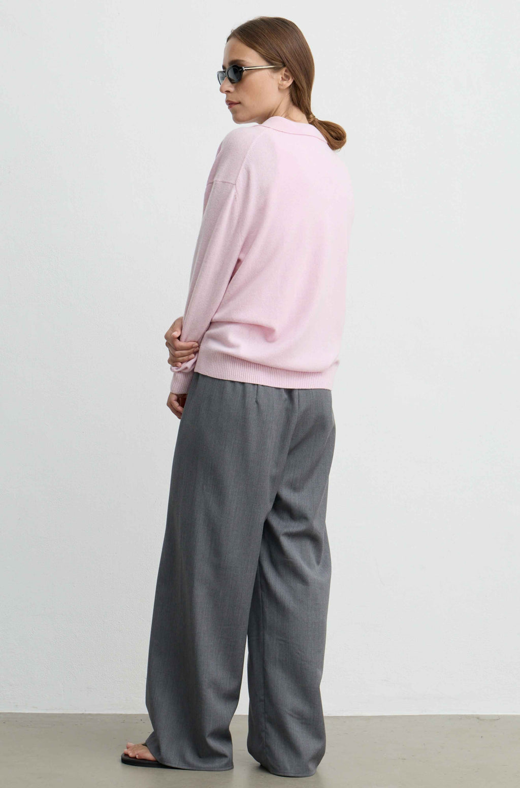 A woman wearing a light pink long-sleeve sweatshirt and wide-leg gray trousers, photographed from the back against a plain light gray studio background.