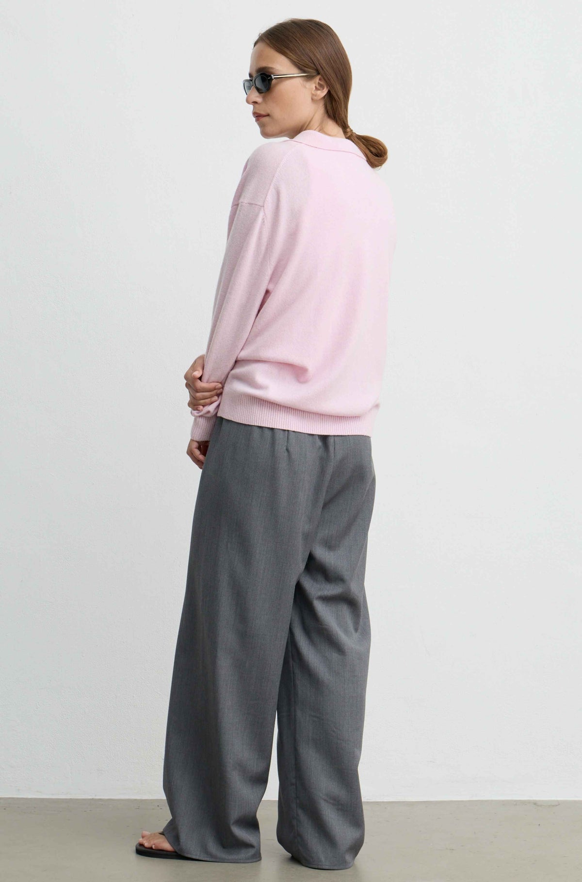 A woman wearing a light pink long-sleeve sweatshirt and wide-leg gray trousers, photographed from the back against a plain light gray studio background.