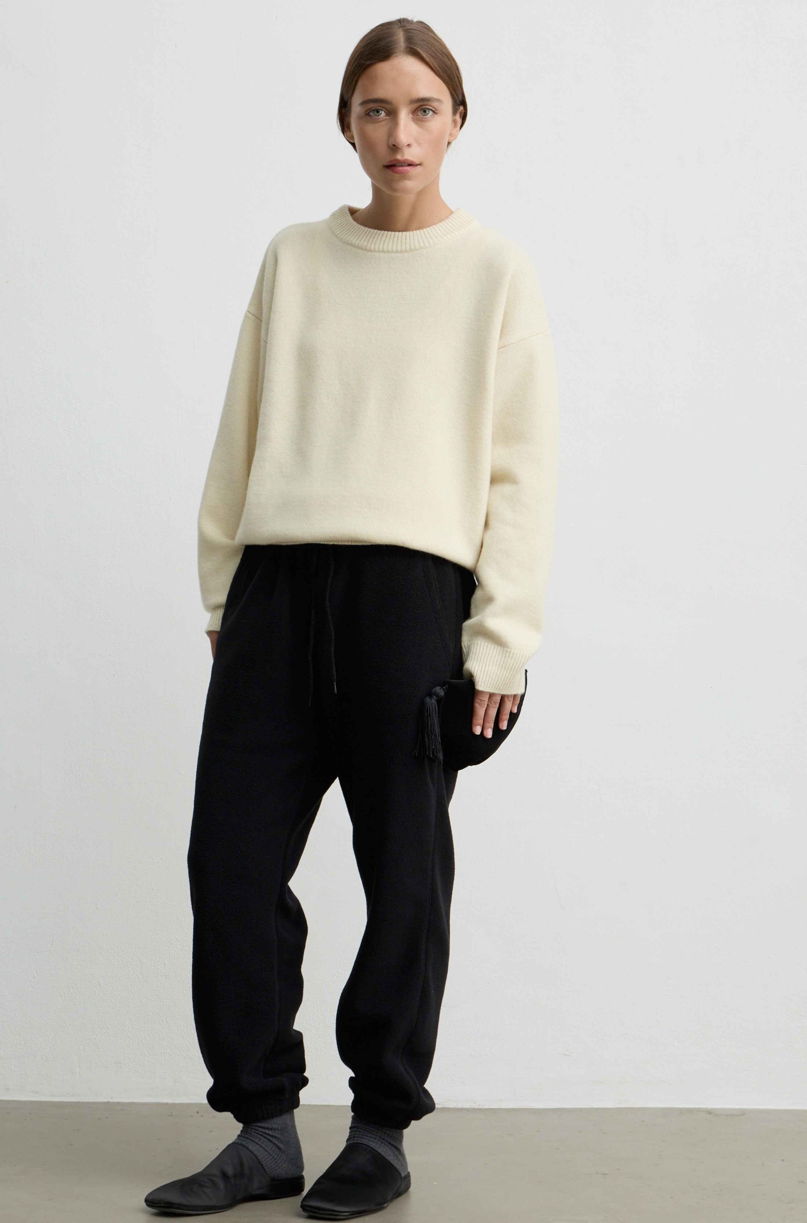 A model stands against a plain light gray studio wall wearing a yellow crewneck sweater, black jogger pants, and dark slip-on shoes.
