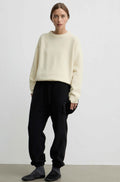 A model stands against a plain light gray studio wall wearing a yellow-colored crewneck sweater, black jogger pants, and dark slip-on shoes.