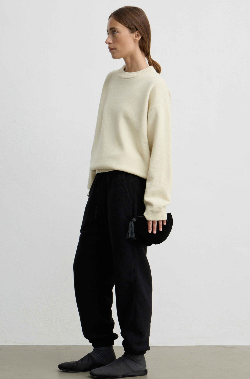 A woman stands in profile wearing a yellow knit sweater, black jogger pants, and black slip-on shoes, holding a small black bag against a plain light gray studio background.