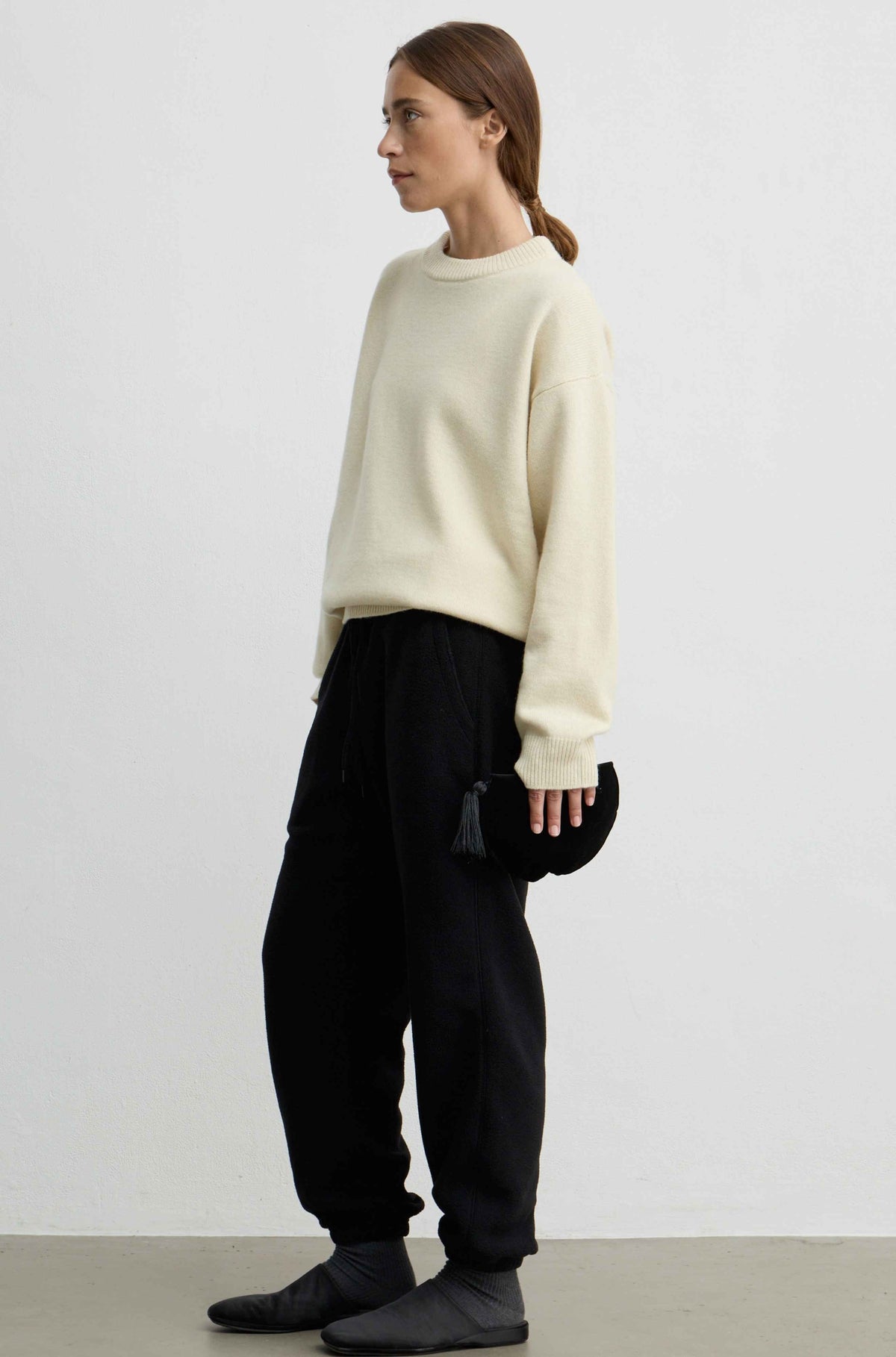 A woman stands in profile wearing a yellow knit sweater, black jogger pants, and black slip-on shoes, holding a small black bag against a plain light gray studio background.