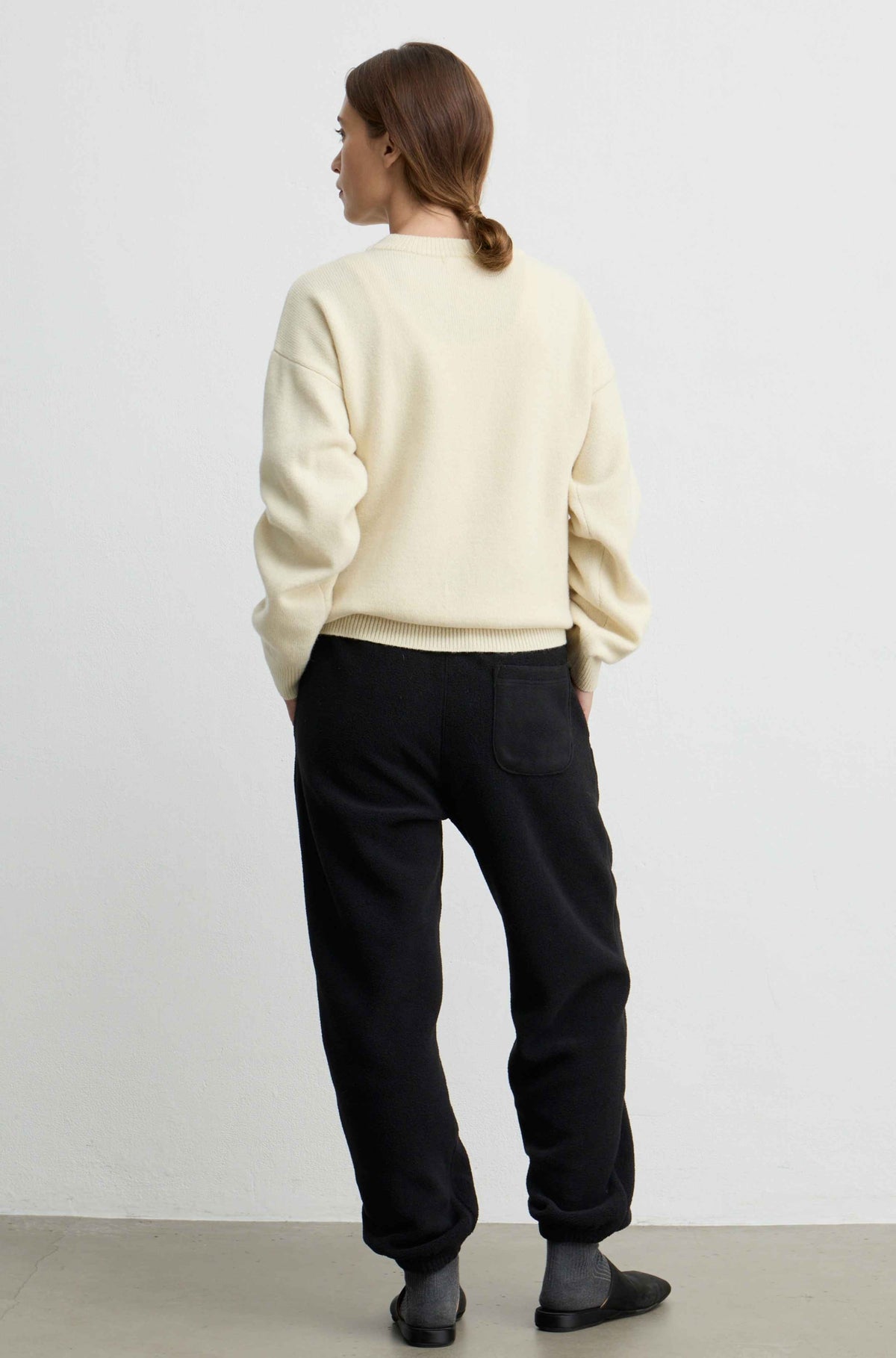 A woman is shown from behind wearing a yellow colored crewneck sweatshirt and black sweatpants against a plain white studio background.
