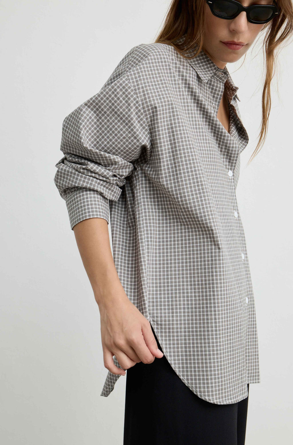 A woman wearing an oversized black and white gingham button-up shirt with rolled-up sleeves, paired with black pants and black sunglasses, photographed against a light gray studio background.
