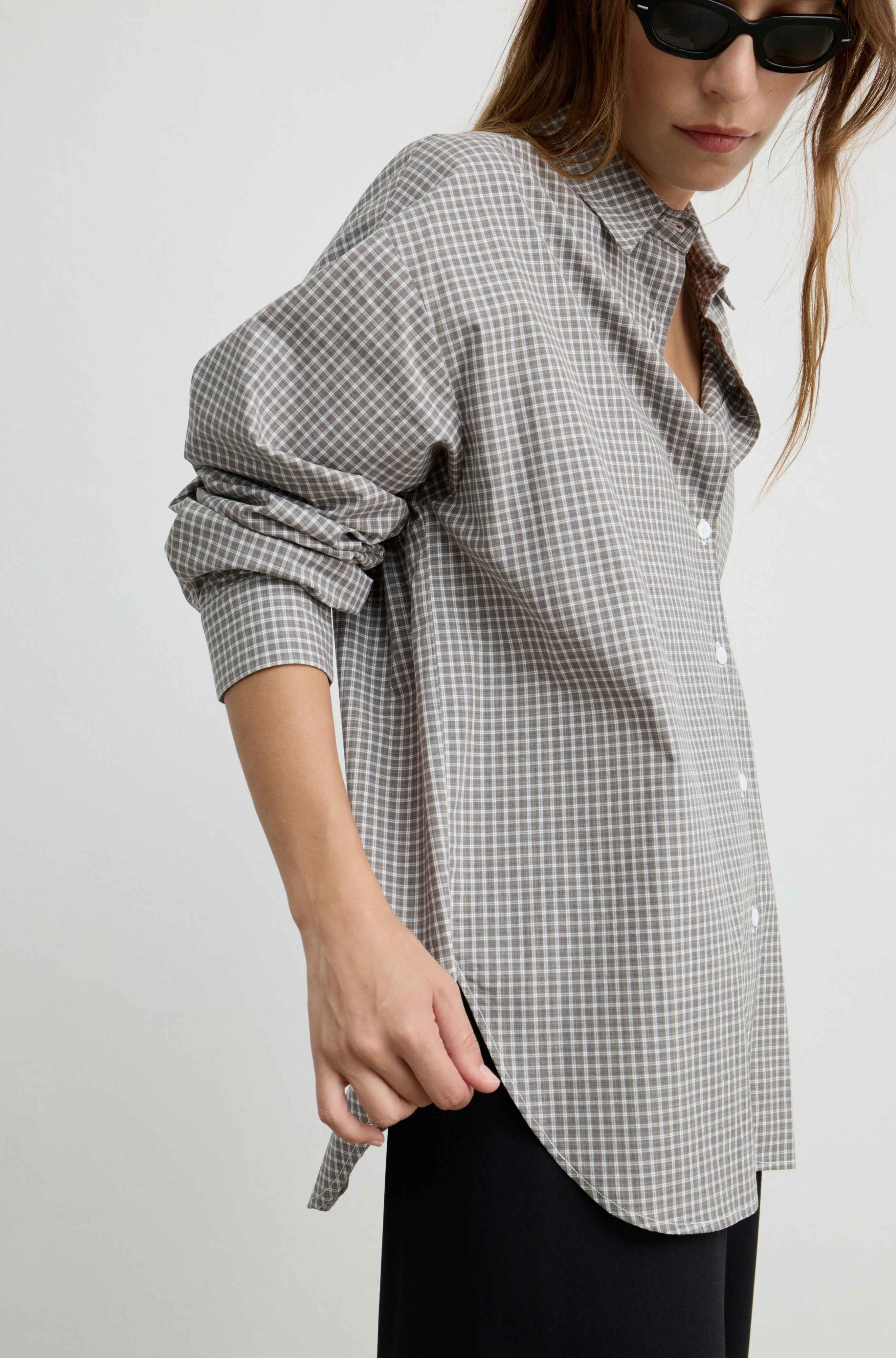 A woman wearing an oversized black and white gingham button-up shirt with rolled-up sleeves, paired with black pants and black sunglasses, photographed against a light gray studio background.