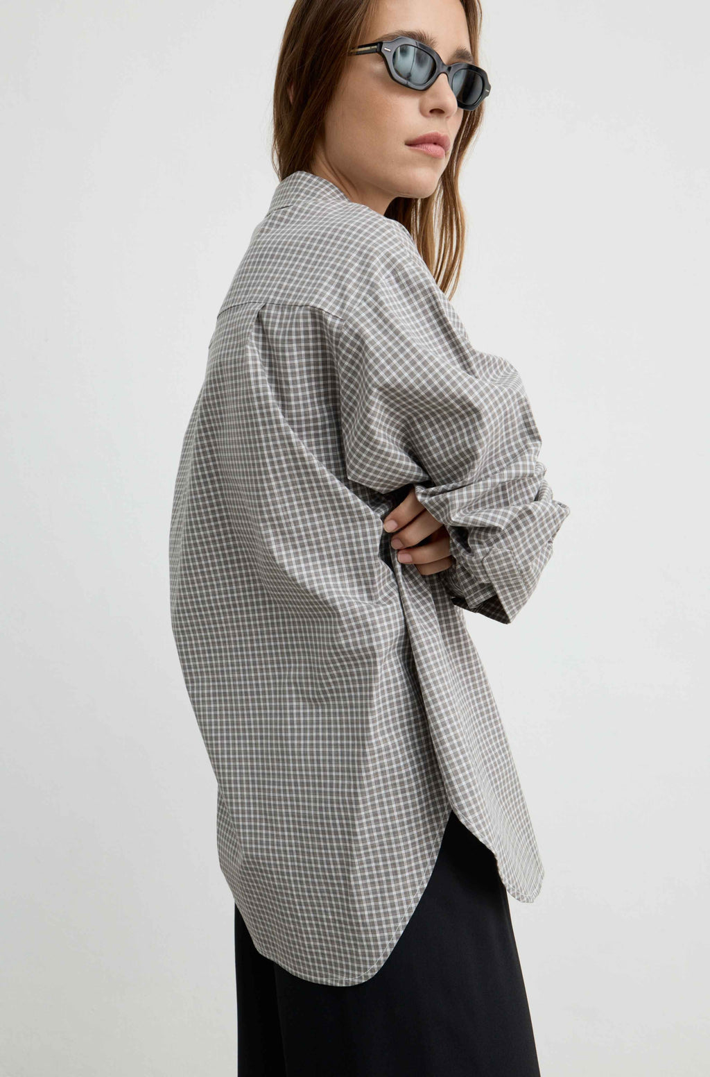 A woman wearing an oversized black and white gingham check shirt, styled with dark sunglasses and a black skirt, photographed against a plain light gray studio background.